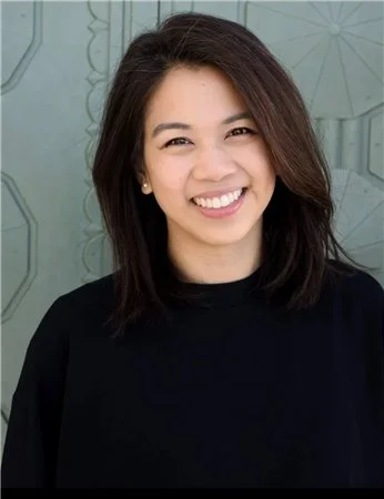 A woman with shoulder-length dark hair smiling confidently in front of a light-colored background with faint geometric patterns. She has a warm caring smile. Easy to approach.