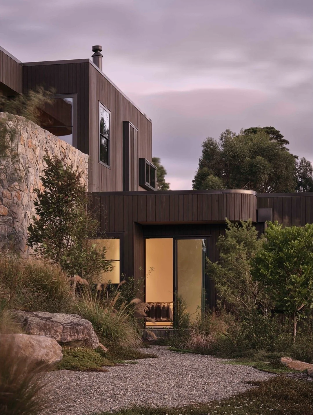 Pink sunset clouds over curved parapet capping, mixed stonework and landscaping at Ti Point House, a Lindesay / Construction Crosson Architects / Rochford Landscapes project where Swift Roofing installed RT7 Roof in ColorCote Horoeka coloured MagnaFl