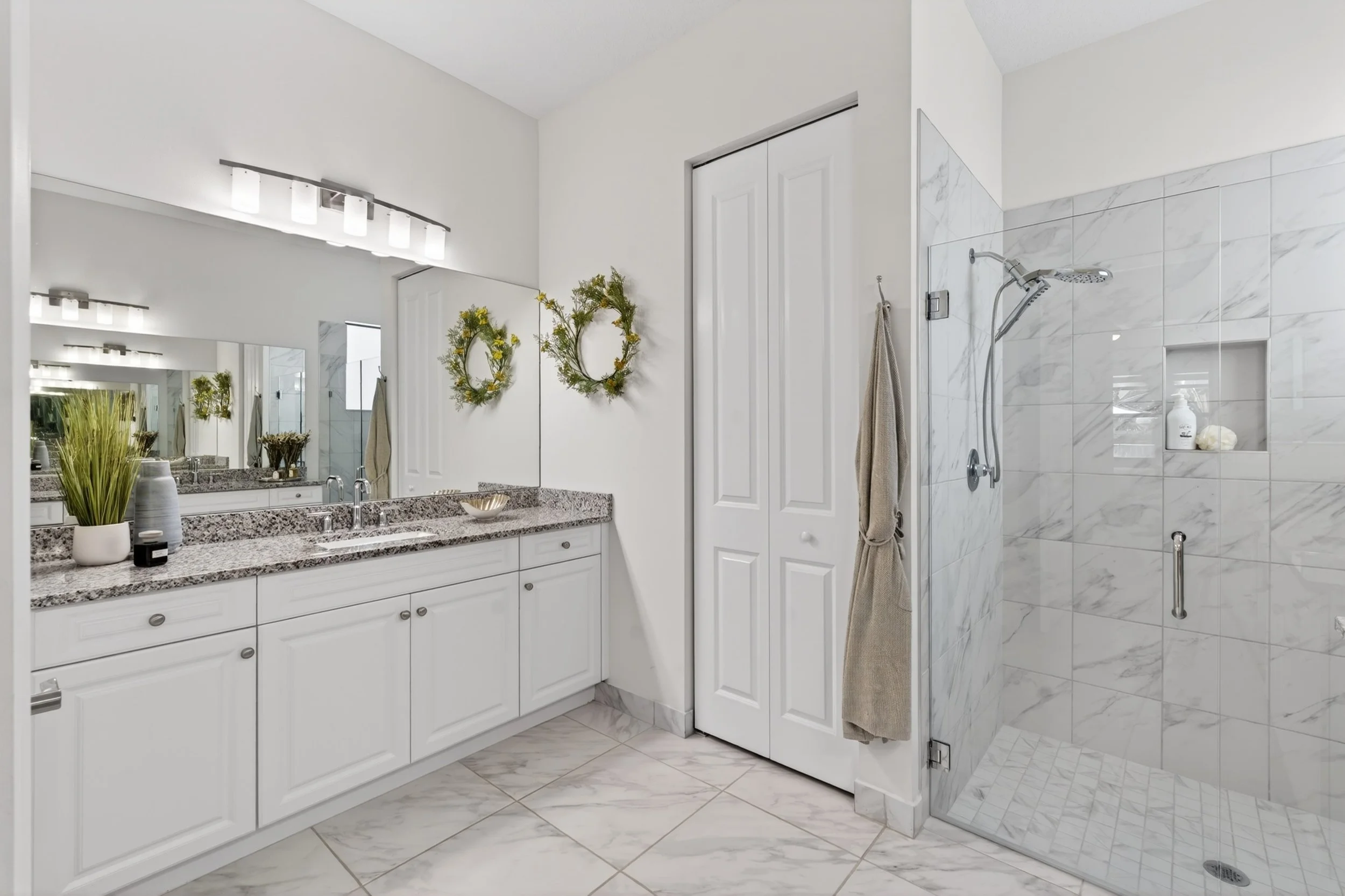 A clean, modern bathroom with a white vanity, granite countertop, decorative plants, large mirror, overhead lighting, and a glass-enclosed shower with marble tiles and a niche for toiletries.