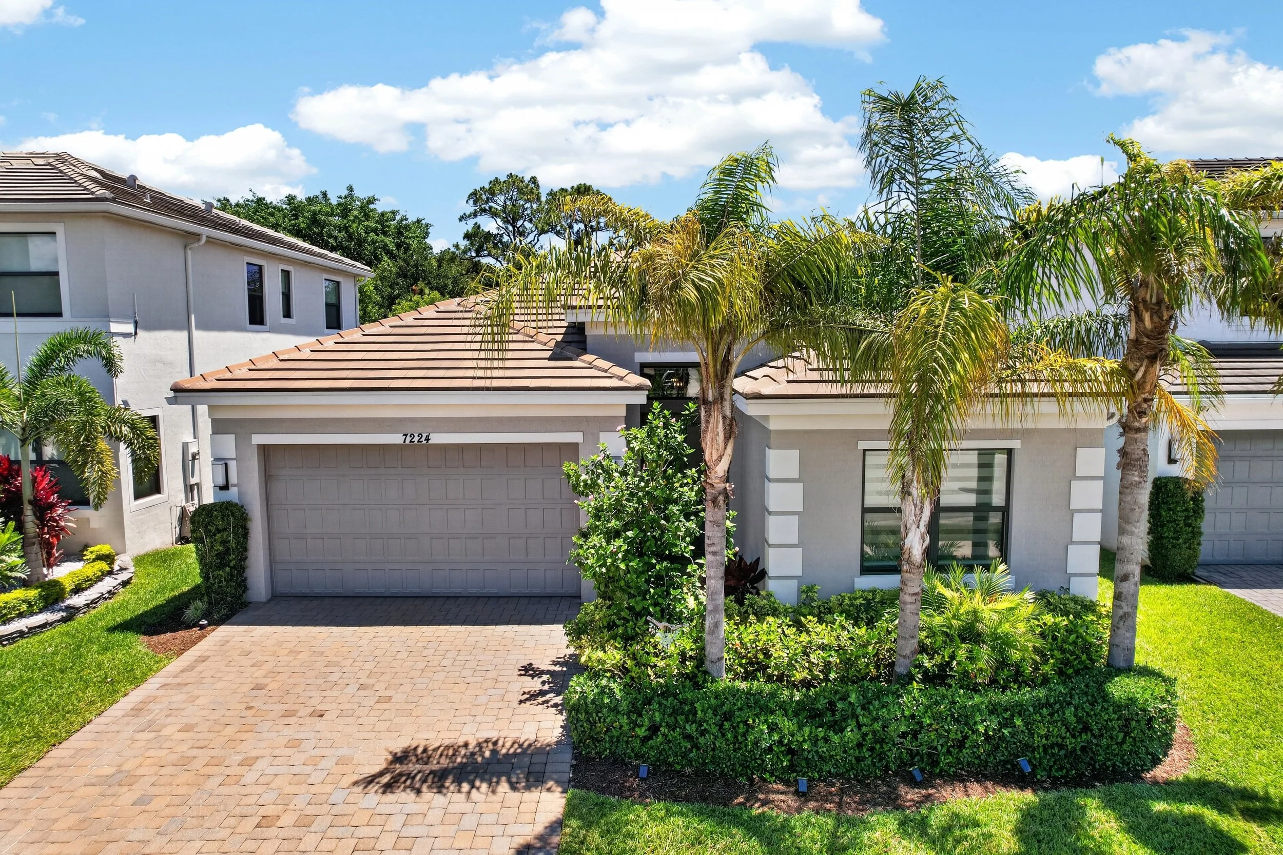Front view of a modern house with a driveway, lush green lawn, flowering bushes, and palm trees under a partly cloudy sky.