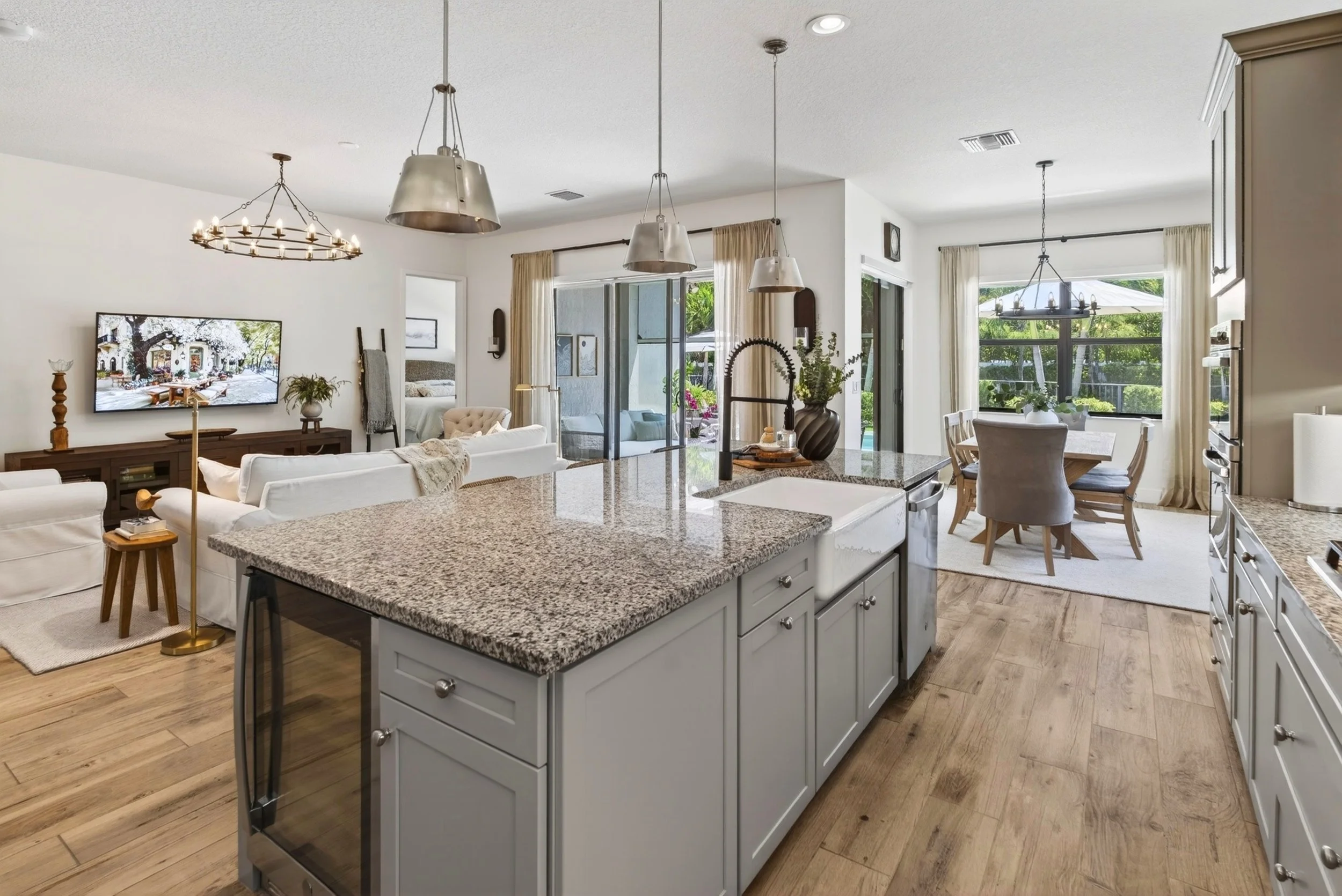 A spacious open-plan living area with a kitchen, dining, and sitting section. The kitchen features a granite countertop island, white cabinets, and a modern sink. The living area has a white sofa, a wall-mounted television, and a beige armchair. Larg