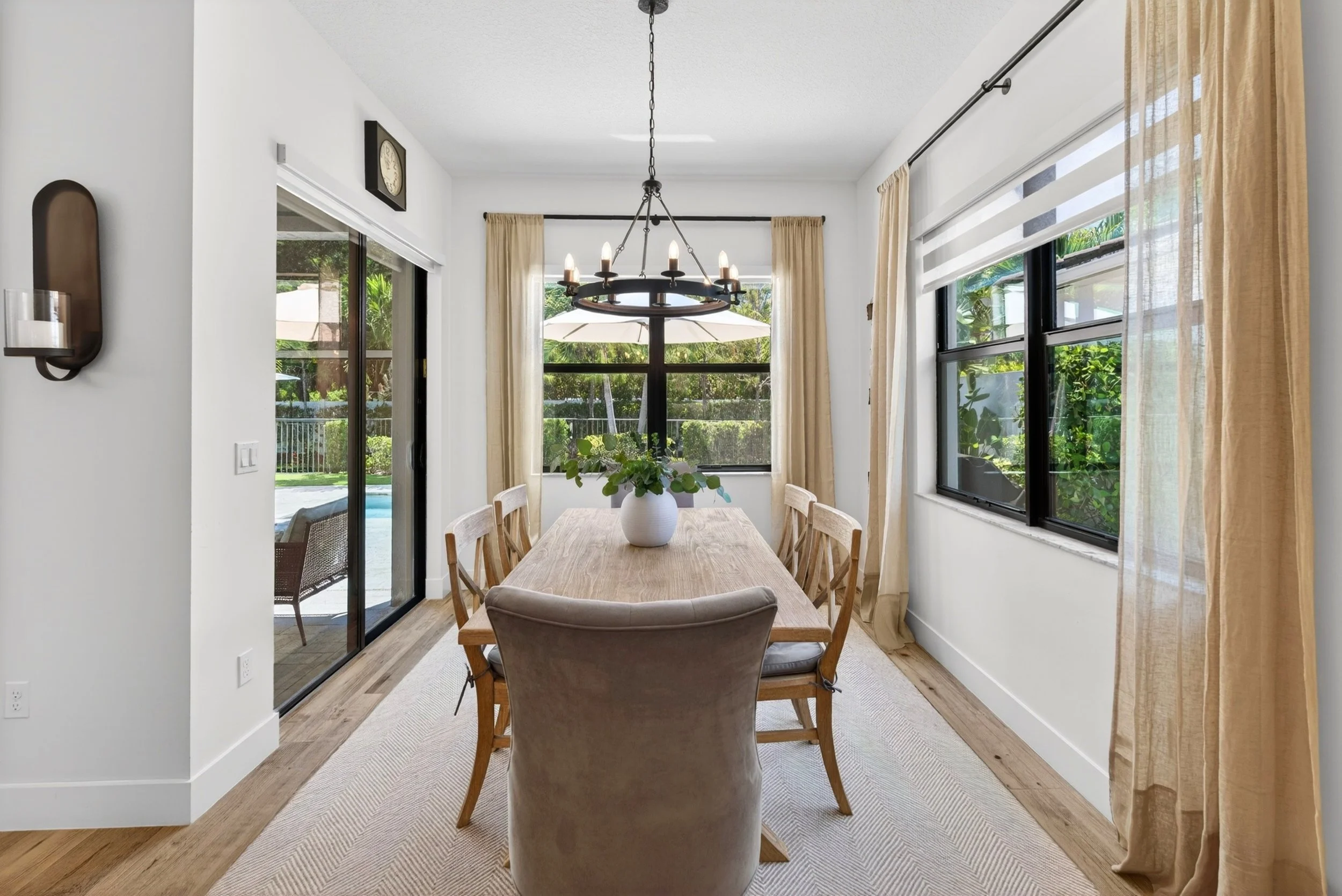 A dining room with a wooden table and six chairs, a white vase with green plants, large windows with cream curtains, a black chandelier hanging from the ceiling, and a view of a backyard with a pool and patio furniture.