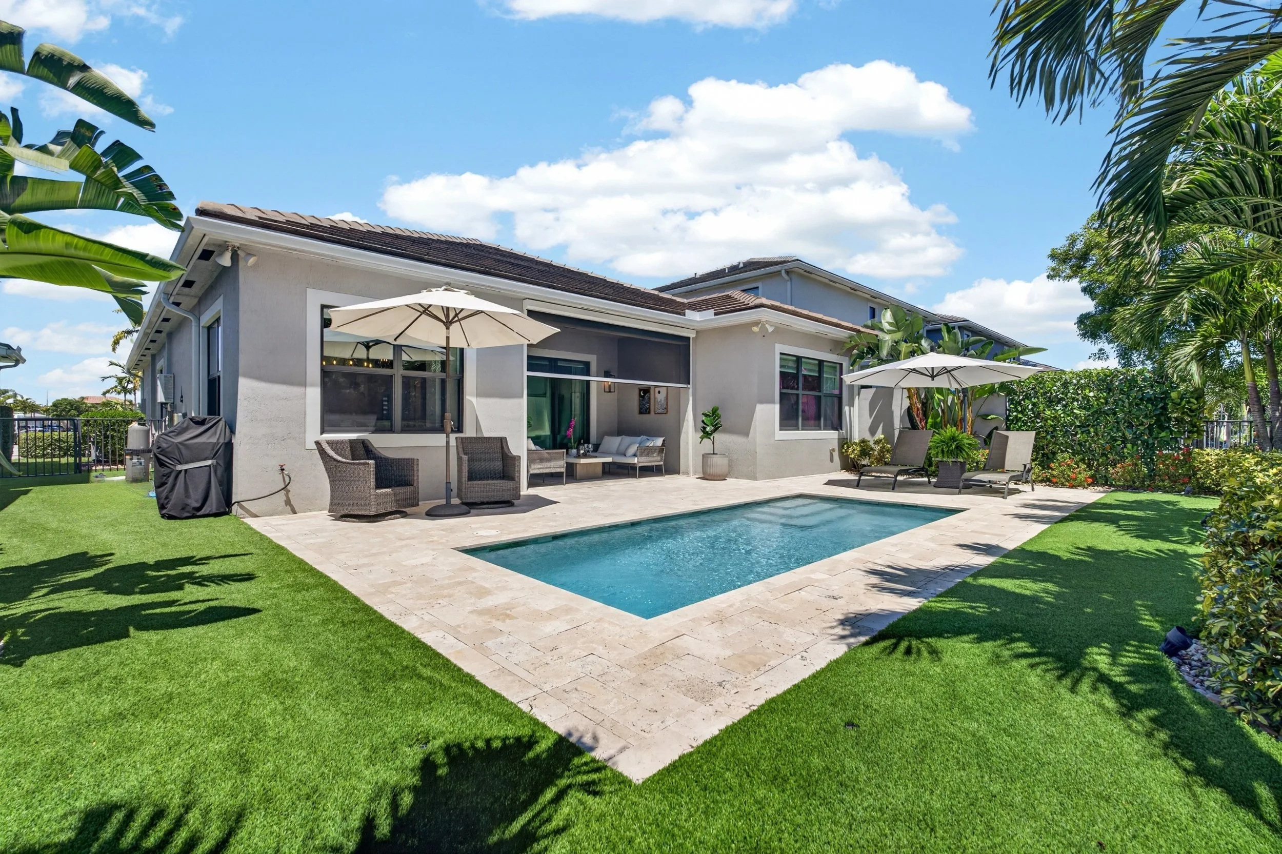 Modern backyard with a small in-ground pool, outdoor seating, umbrellas, and lush green grass, with a house and trees in the background under a blue sky with clouds.
