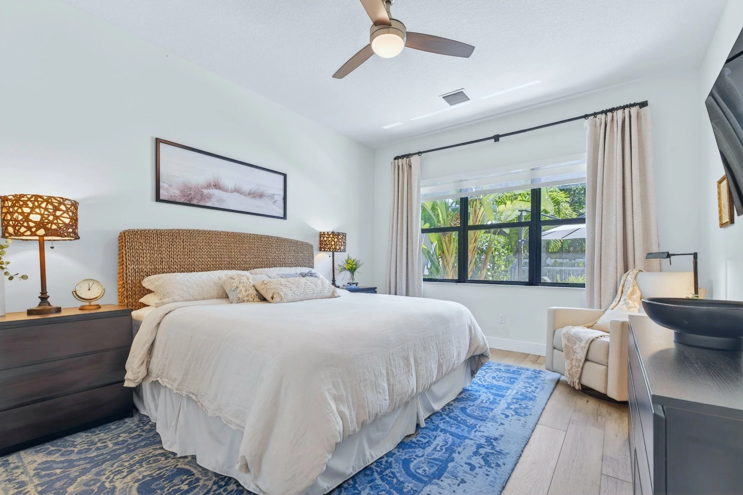 A bedroom with a large bed, two nightstands with lamps, a framed picture on the wall, a window with curtains and a view of greenery outside, and a chair in the corner.