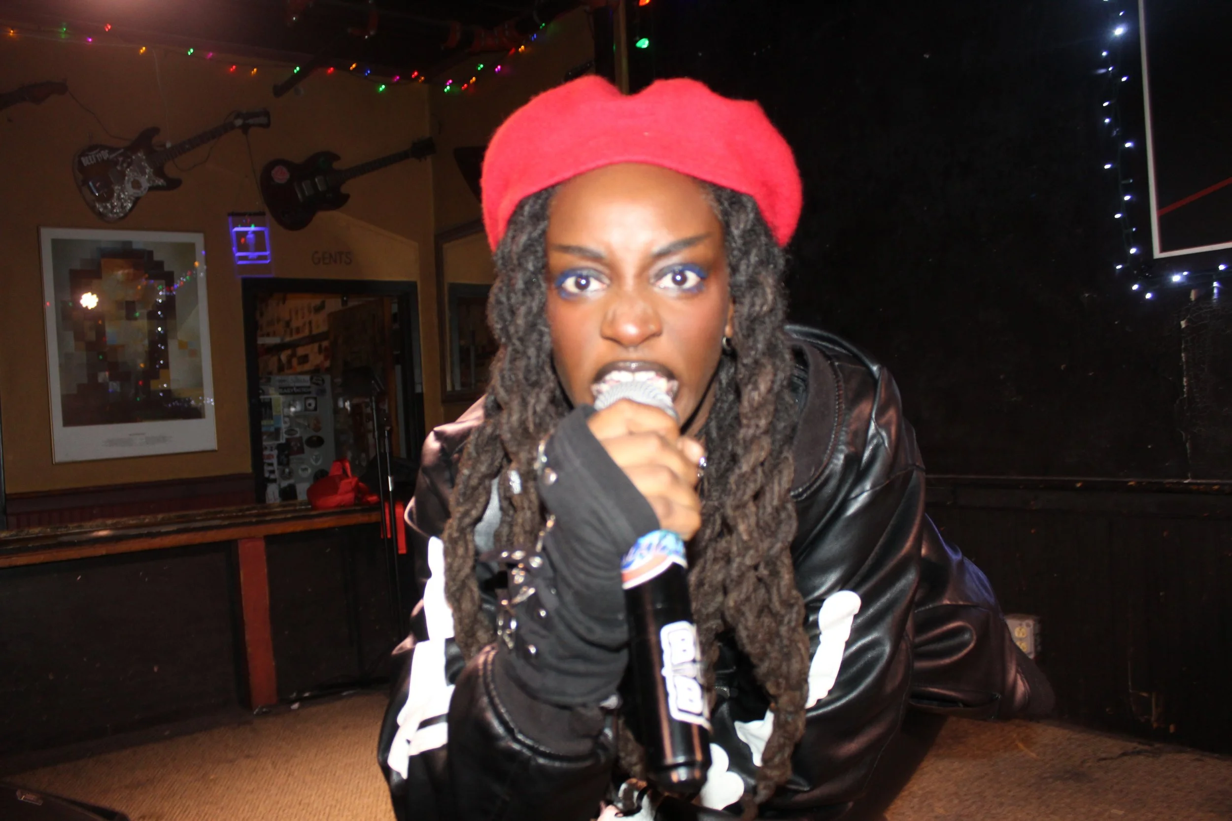 Woman with long dreadlocks wearing a red beret, black leather jacket, and black gloves, holding a microphone on stage at a performance venue decorated with guitars and colorful string lights.