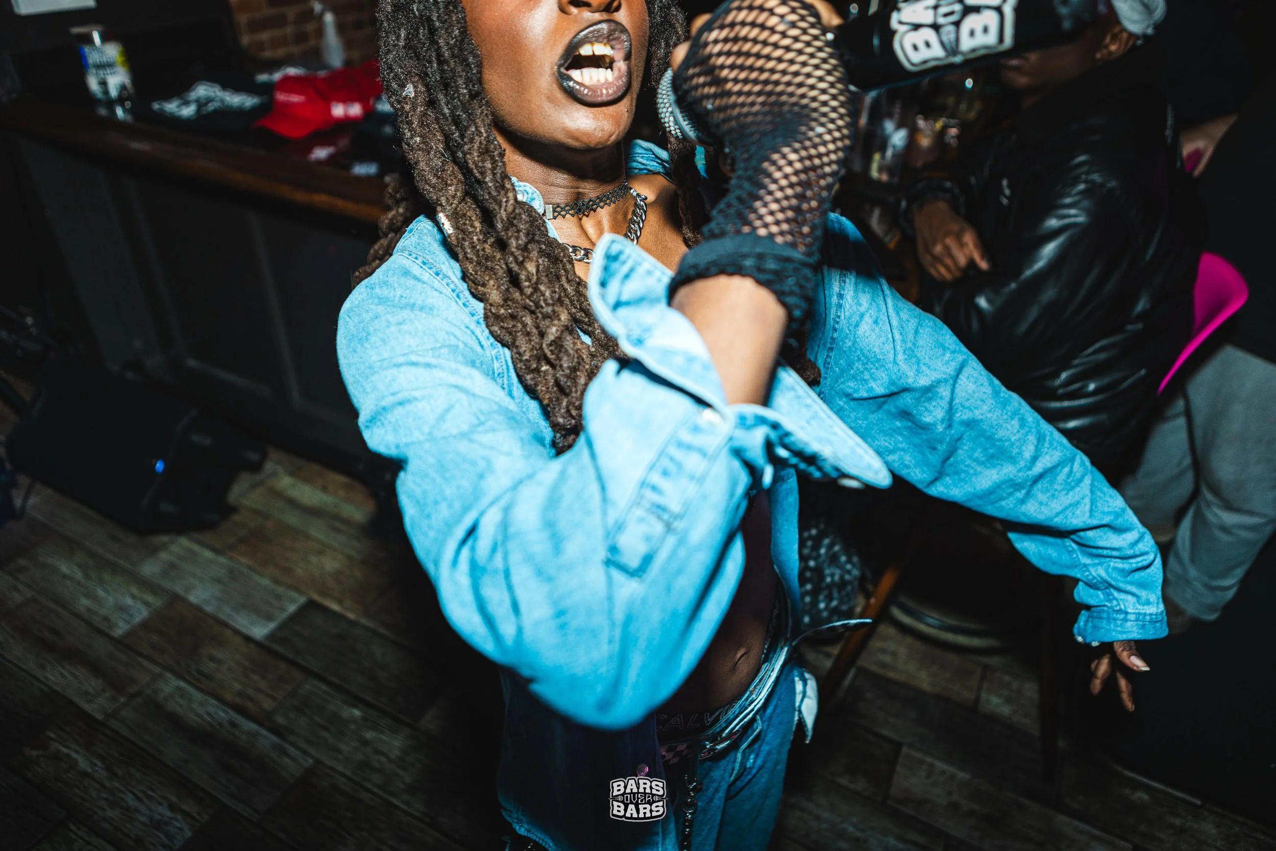 A woman with dreadlocks, Militia Rashad, wearing a denim jacket, is holding a microphone and appears to be raging her f****** heart out.