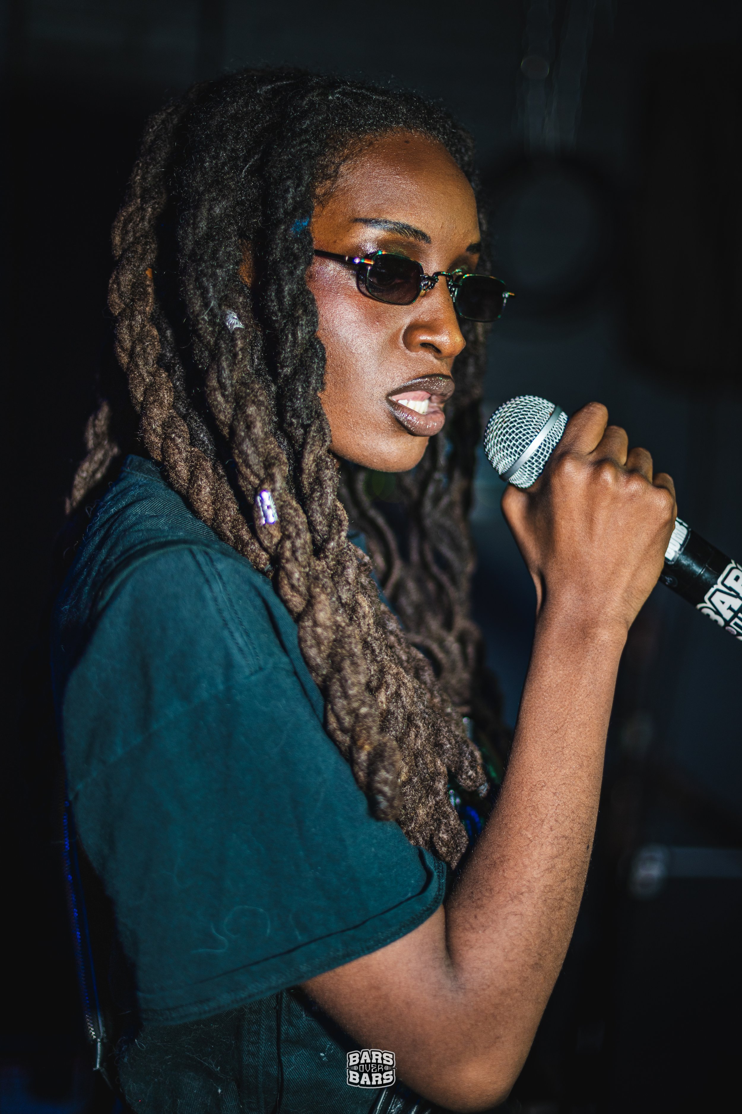 A woman with dreadlocks, wearing sunglasses and a dark shirt, holding a microphone and performing or speaking.