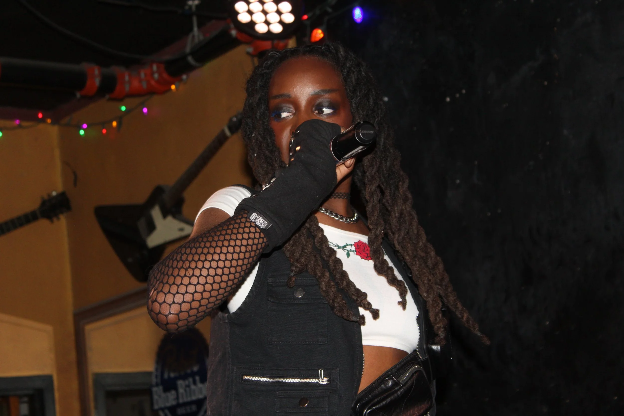 A woman with dreadlocks holding a microphone on stage, wearing a fishnet sleeve glove, a white crop top with a rose embroidery, and a black vest. Dim stage lighting with colorful string lights in the background.