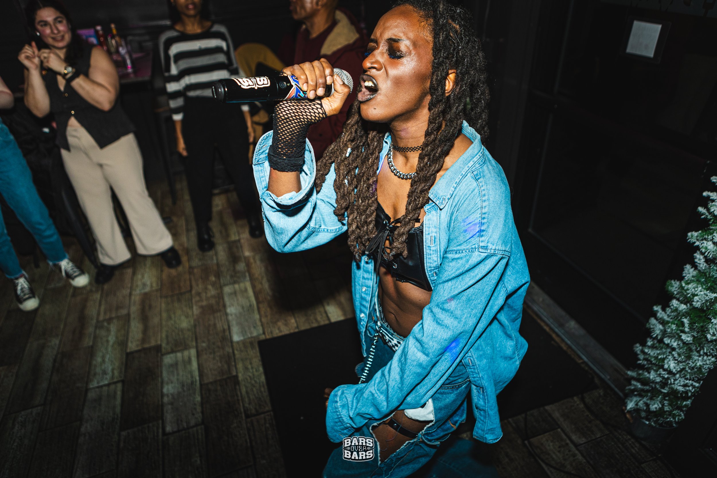 A femme singer with long locs, Militia Rashad, wearing a denim jacket and fishnet gloves, passionately raging into a microphone on stage.