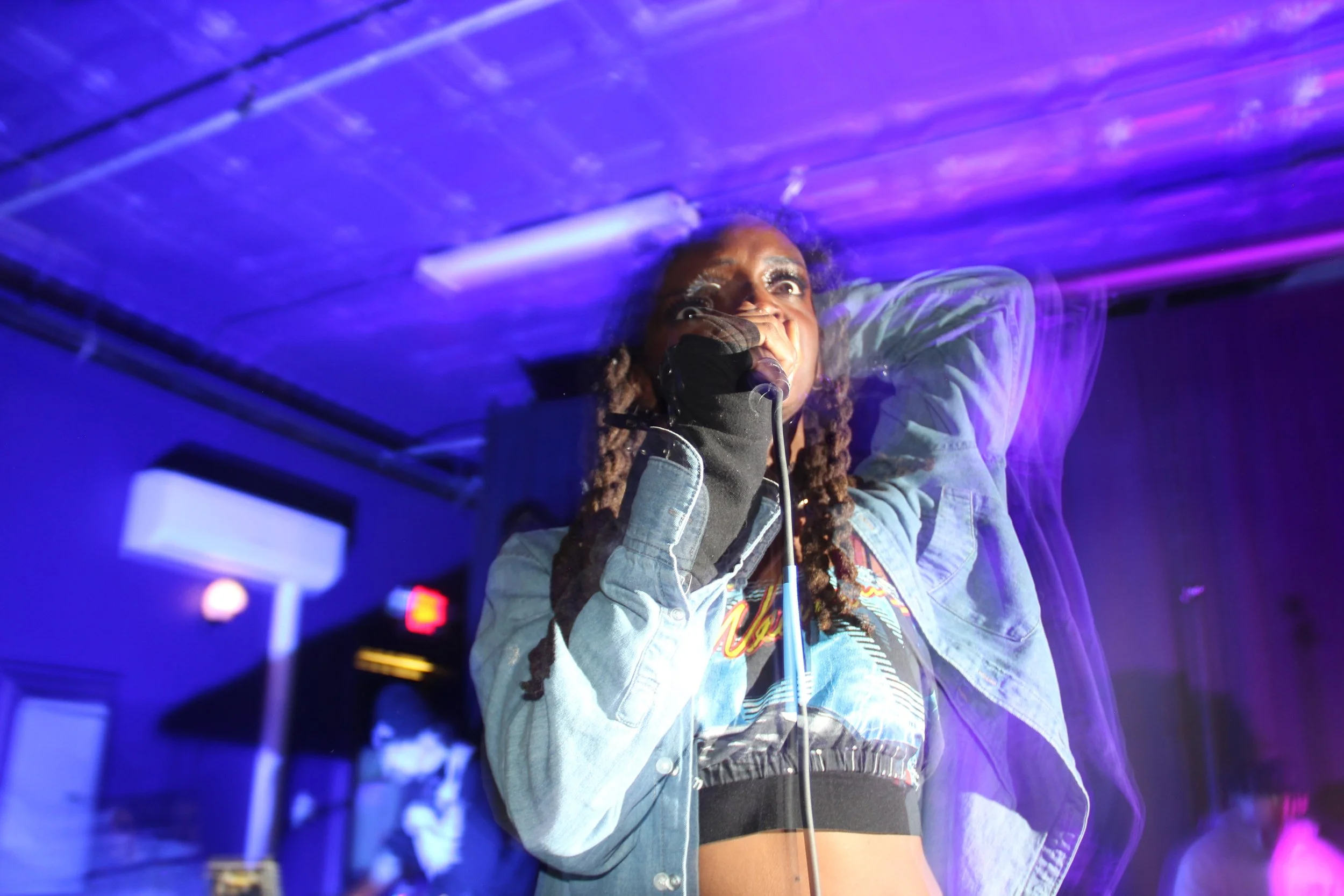 A woman with long dreadlocks wearing a denim jacket and a black crop top performs on stage with purple lighting in the background.