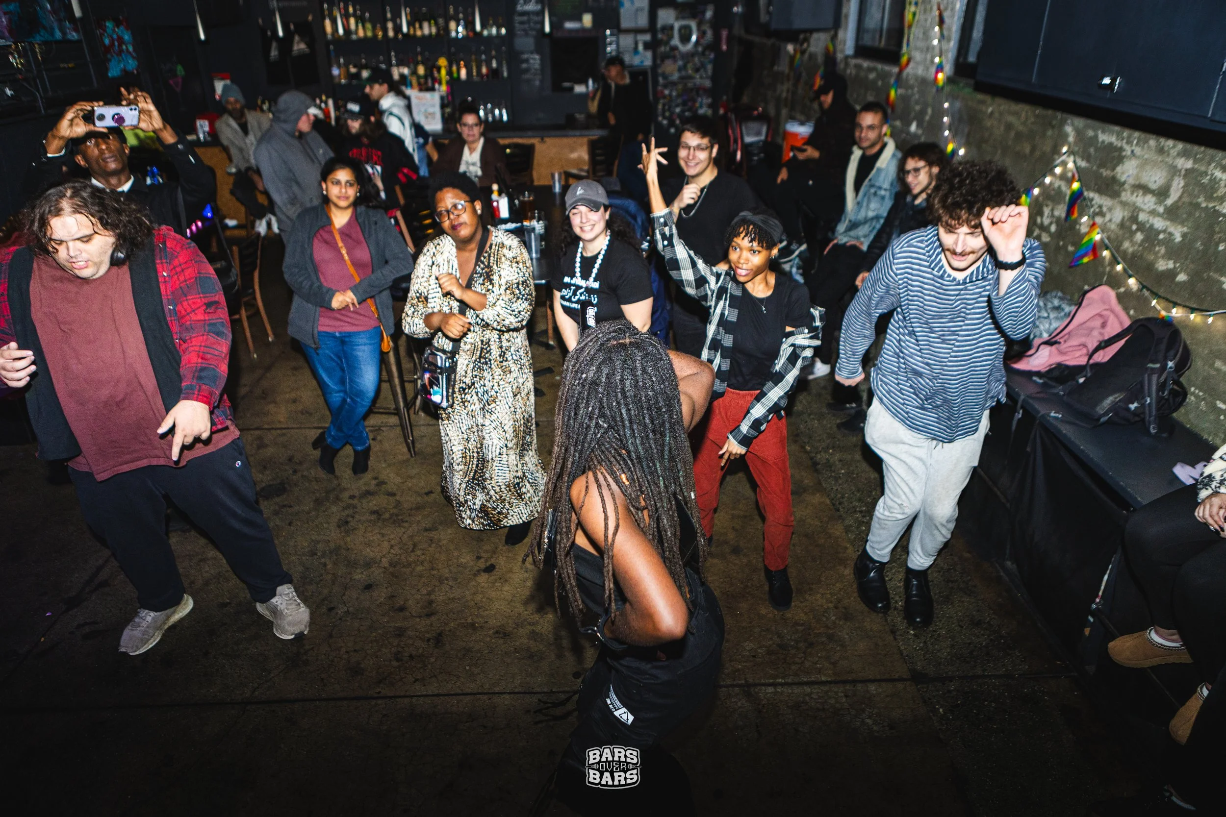 People dancing and socializing at a lively indoor party or club, with colorful decorations and a bar in the background.