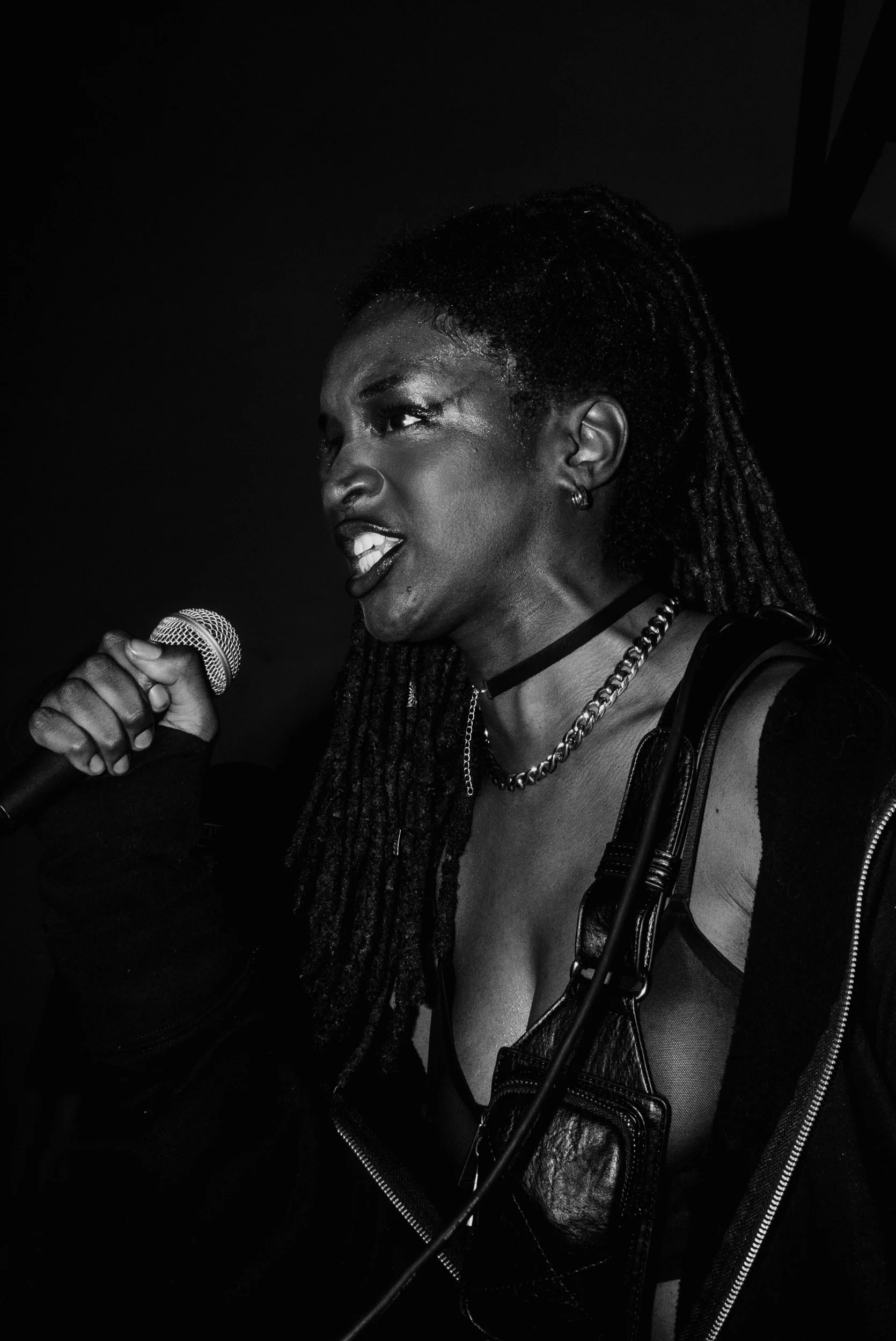 Black and white photo of a woman singing or speaking into a microphone with an intense expression, wearing a leather jacket and chained necklace.