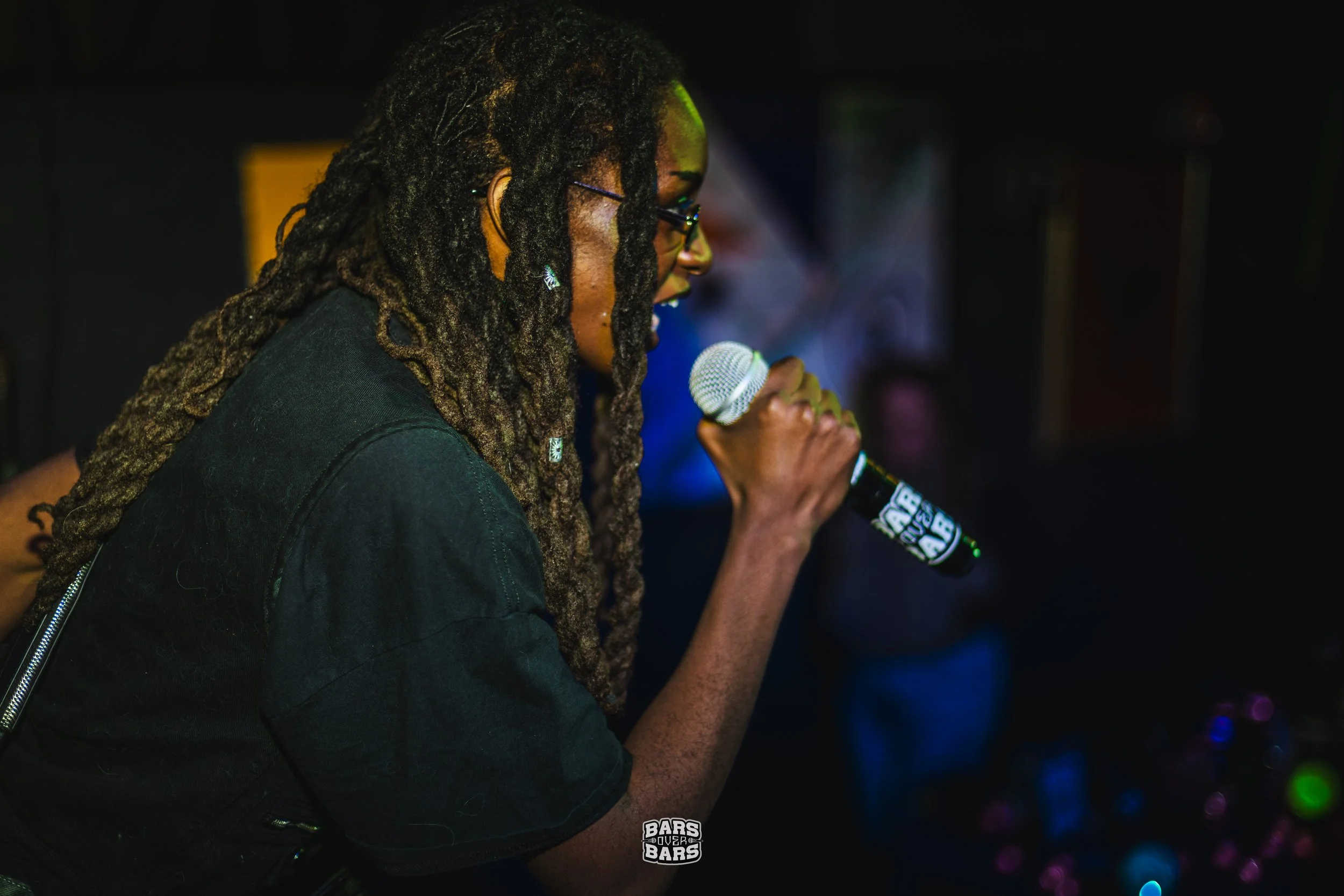 A woman with long dreadlocks and glasses holding a microphone while speaking or singing at a dimly lit event.