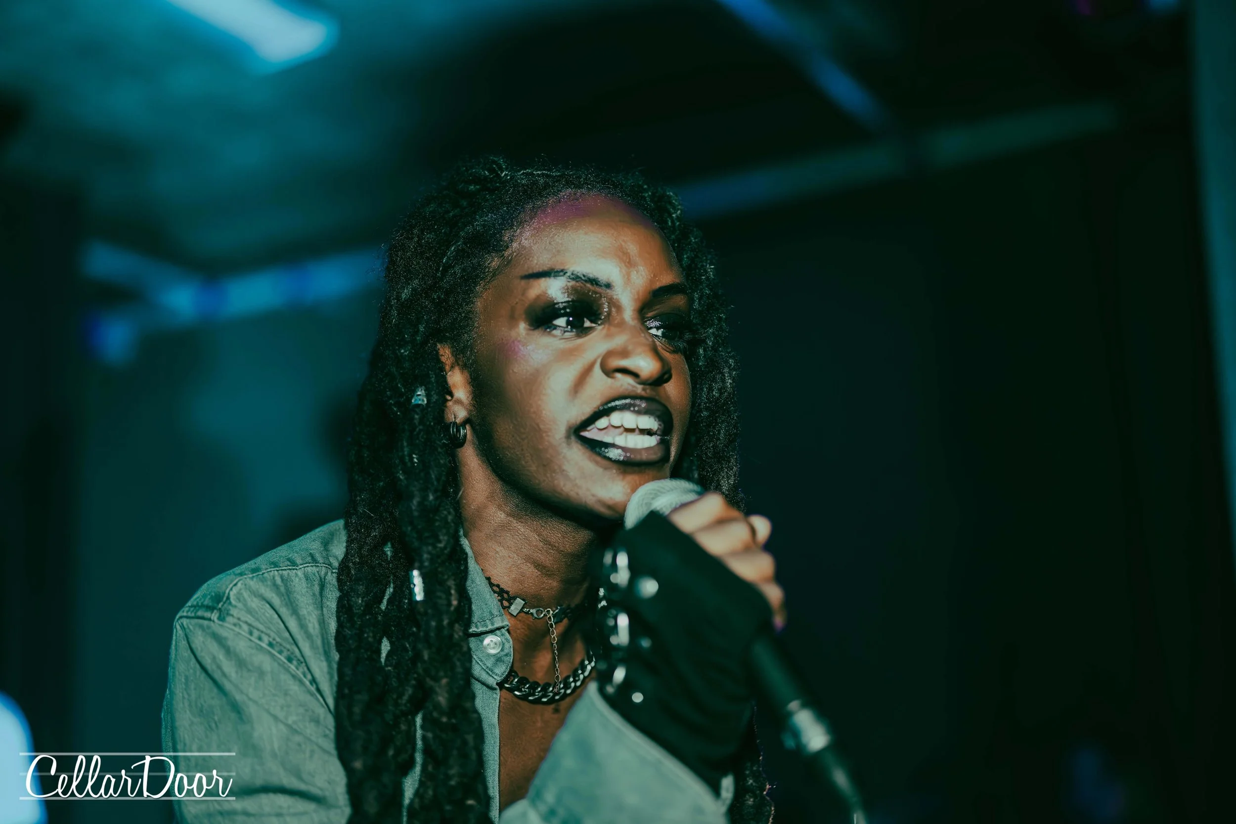 Woman with dreadlocks holding a microphone, speaking passionately at a dimly lit event.
