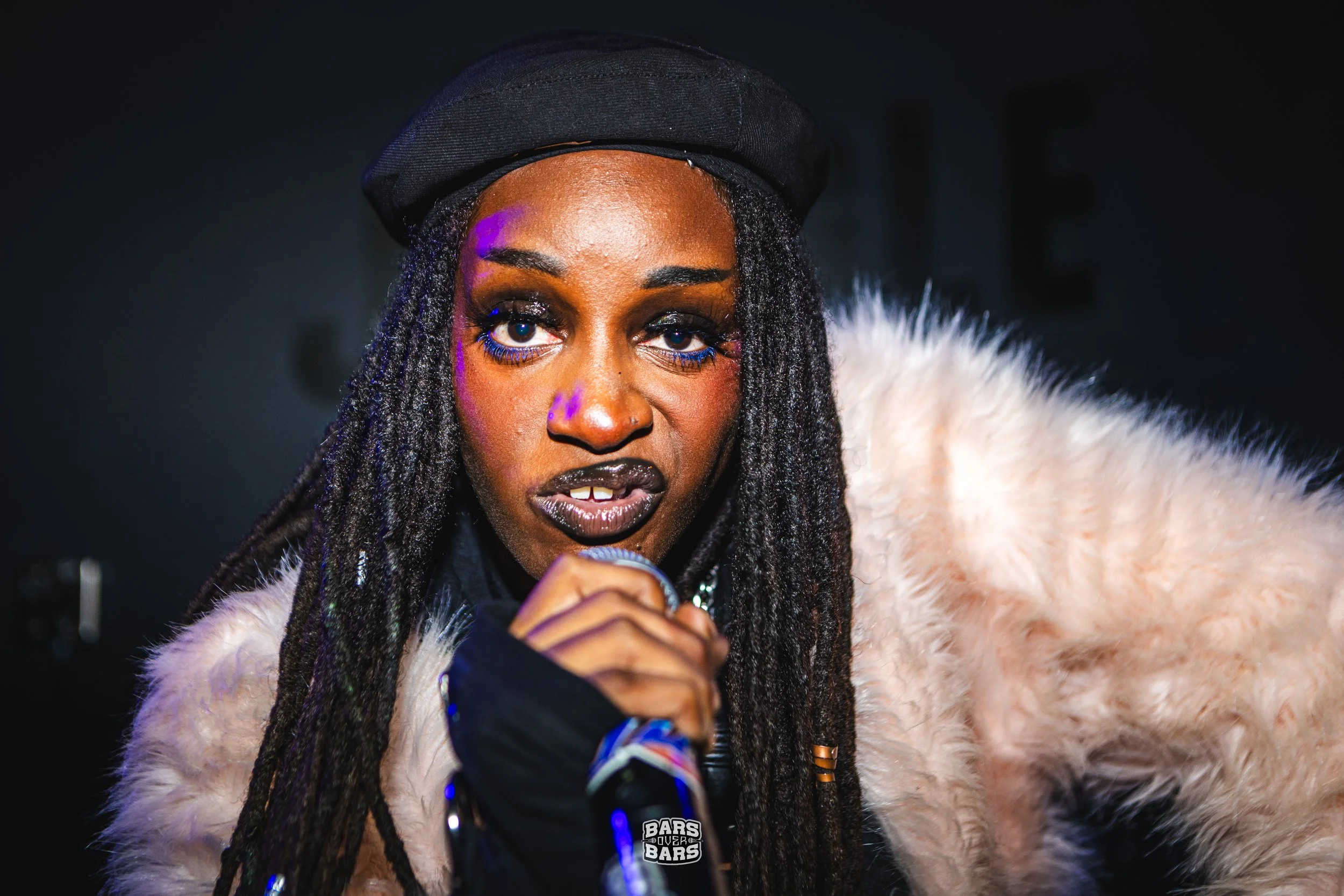 Close-up of a woman with long dreadlocks wearing a black beret, makeup with blue mascara, a finger near her mouth, and a fluffy light-colored garment, holding a microphone.