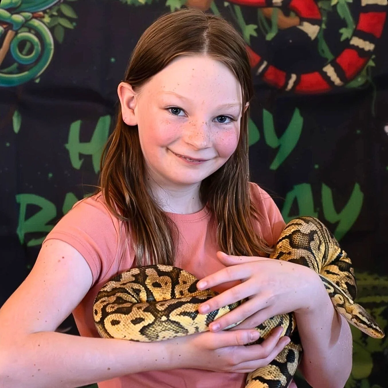 A young girl with long red hair and freckles holding a yellow and brown snake. She is smiling and wearing a pink shirt, standing in front of a colorful backdrop with green and red designs.