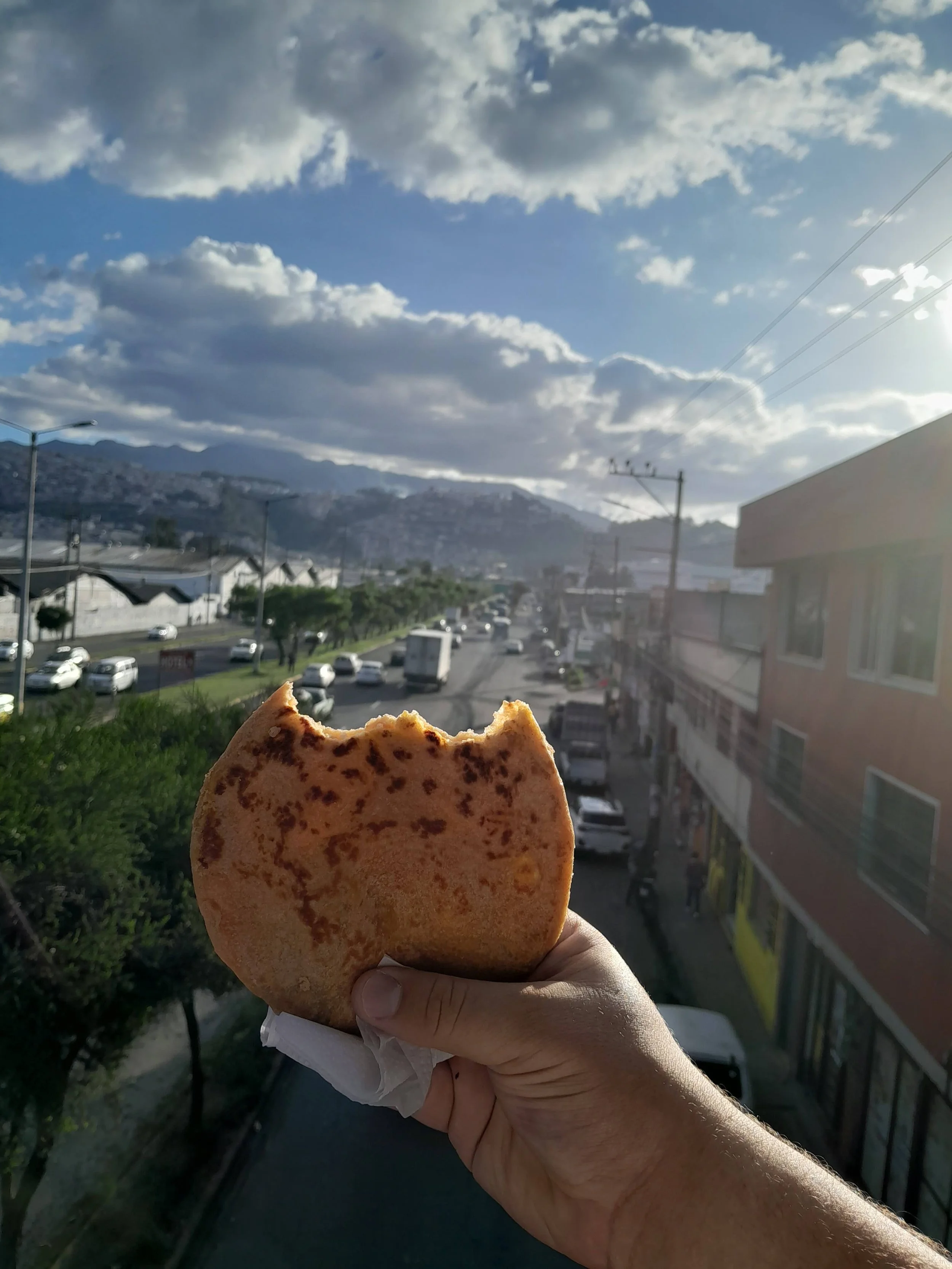 A hand holding a partially eaten breakfast taco with a city street, cars, and mountains in the background during cloudy weather.