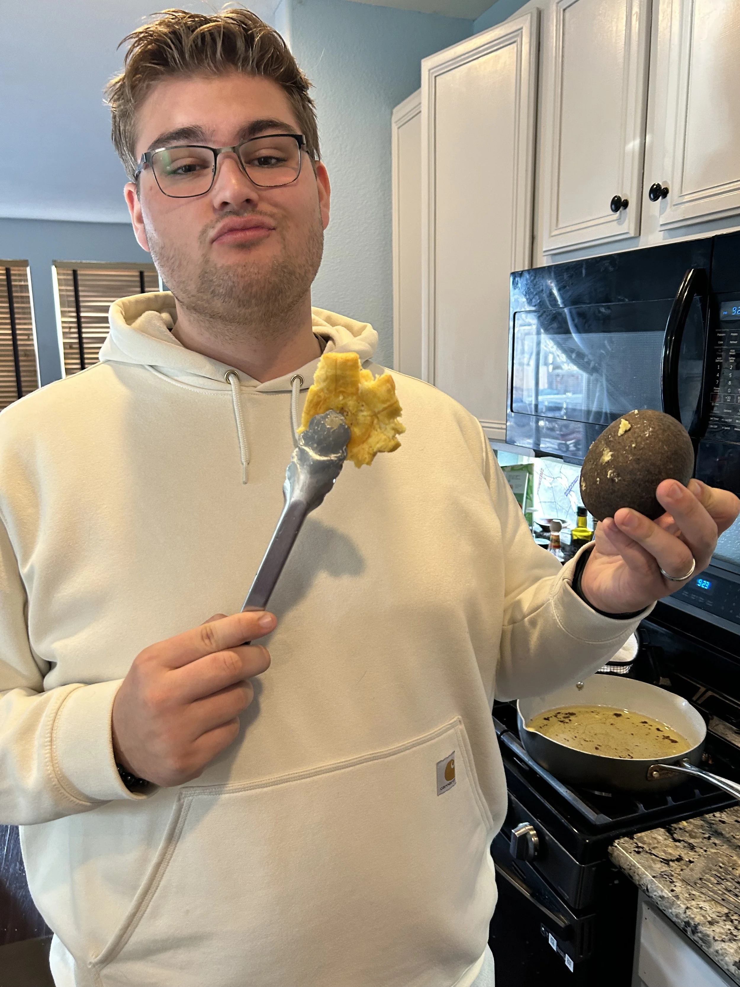 A man in a cream-colored hoodie holding a fork with a piece of food in one hand and a coarse, dark-colored ball-shaped object in the other, standing in a kitchen with white cabinets and a stove with a cooking pan.