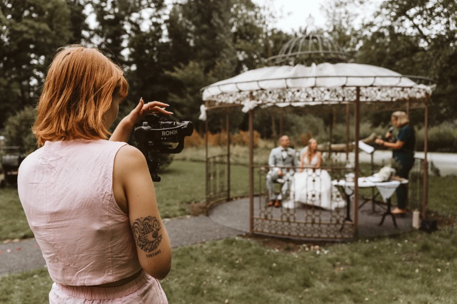 Eine Frau mit roten Haaren und Tattoo auf dem Arm filmt eine Hochzeitszeremonie mit einer Kamera. Im Hintergrund sind ein Brautpaar und ein Zeremonienleiter unter einer dekorativen Pagode zu sehen.