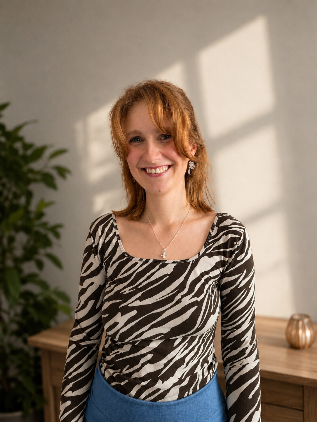 Junge Frau mit roten Haaren, schwarzem und weißem Muster-Shirt, blauer Hose, Haarnadel, Kette mit Buchstaben, steht im Raum mit Schreibtisch und Pflanze, Sonnenlicht auf der Wand.