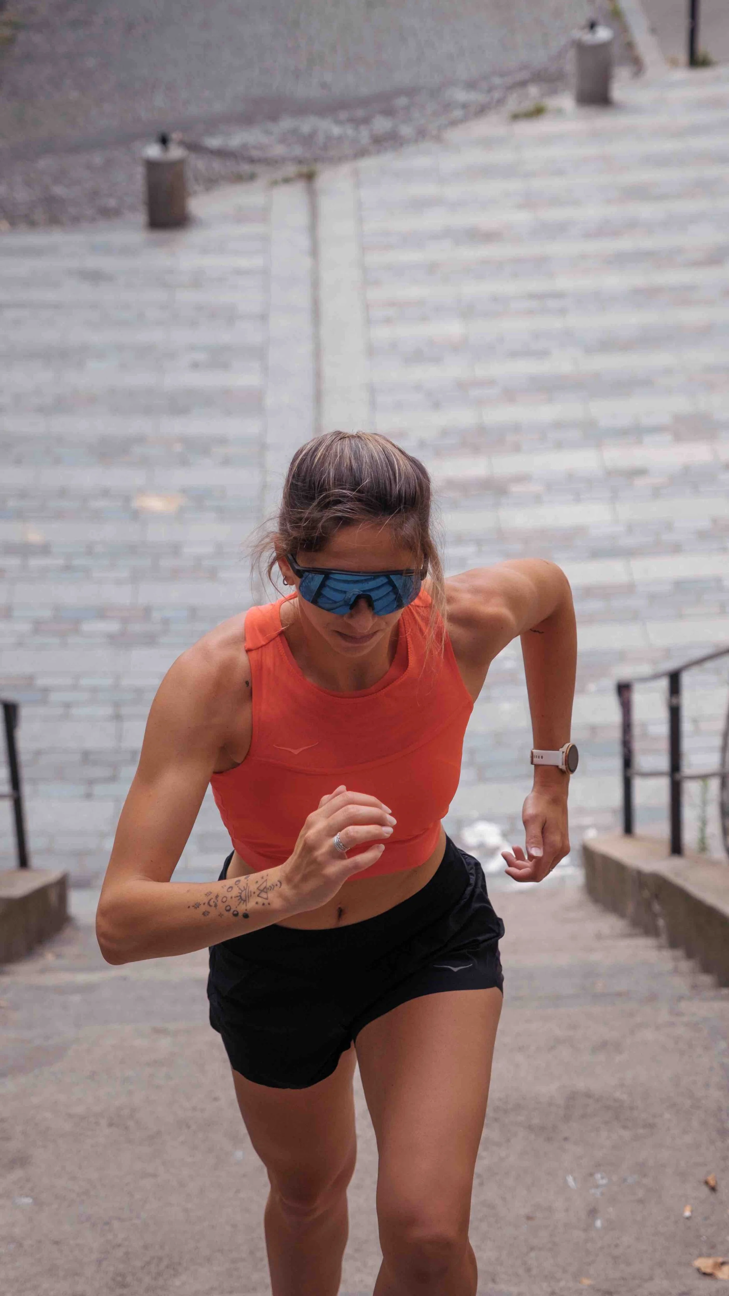 Une femme en tenue de course, portant un vêtement orange et des lunettes de sport, en train de courir en extérieur.