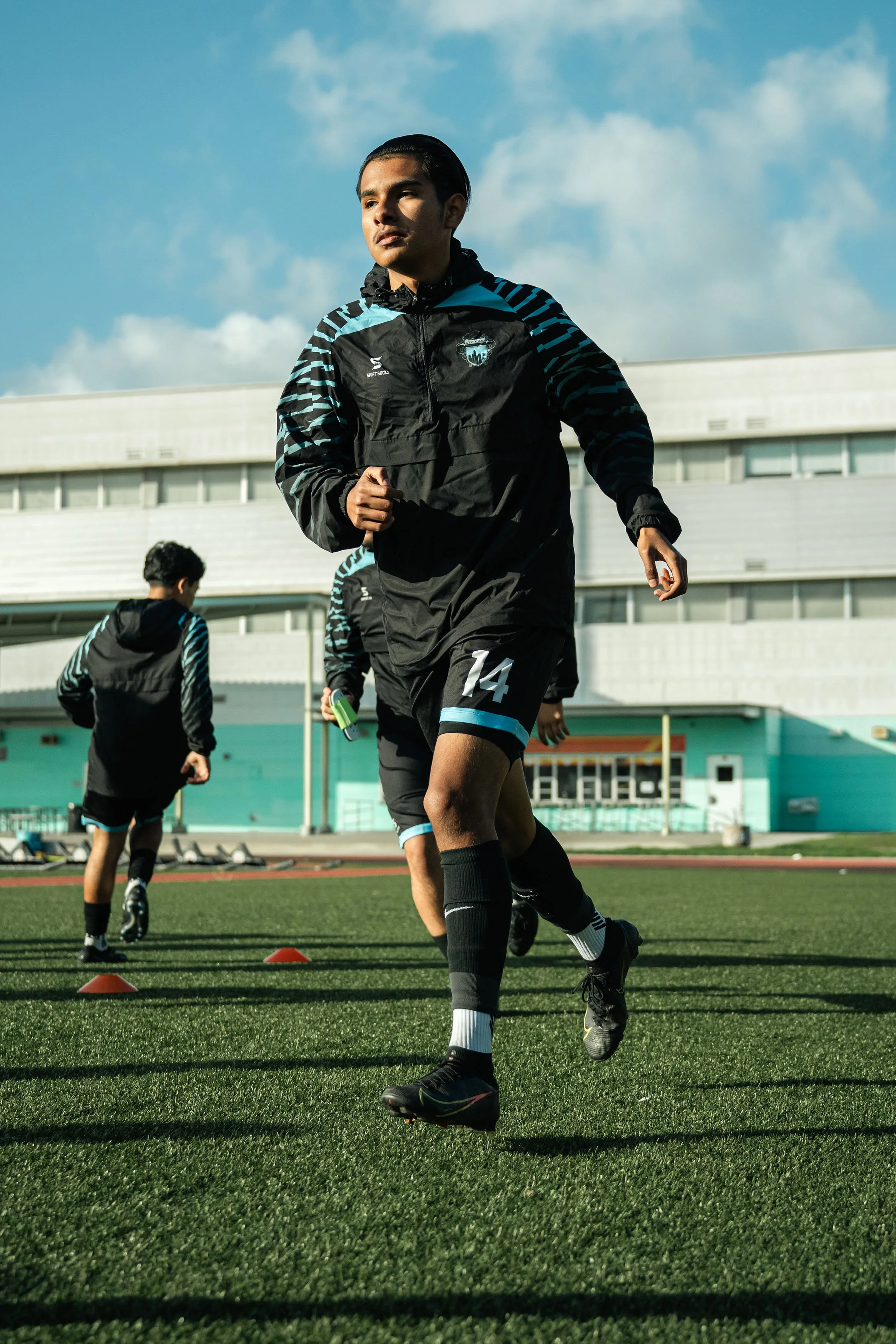 Soccer players in black training attire jogging on a sports field with cones, under a blue sky.
