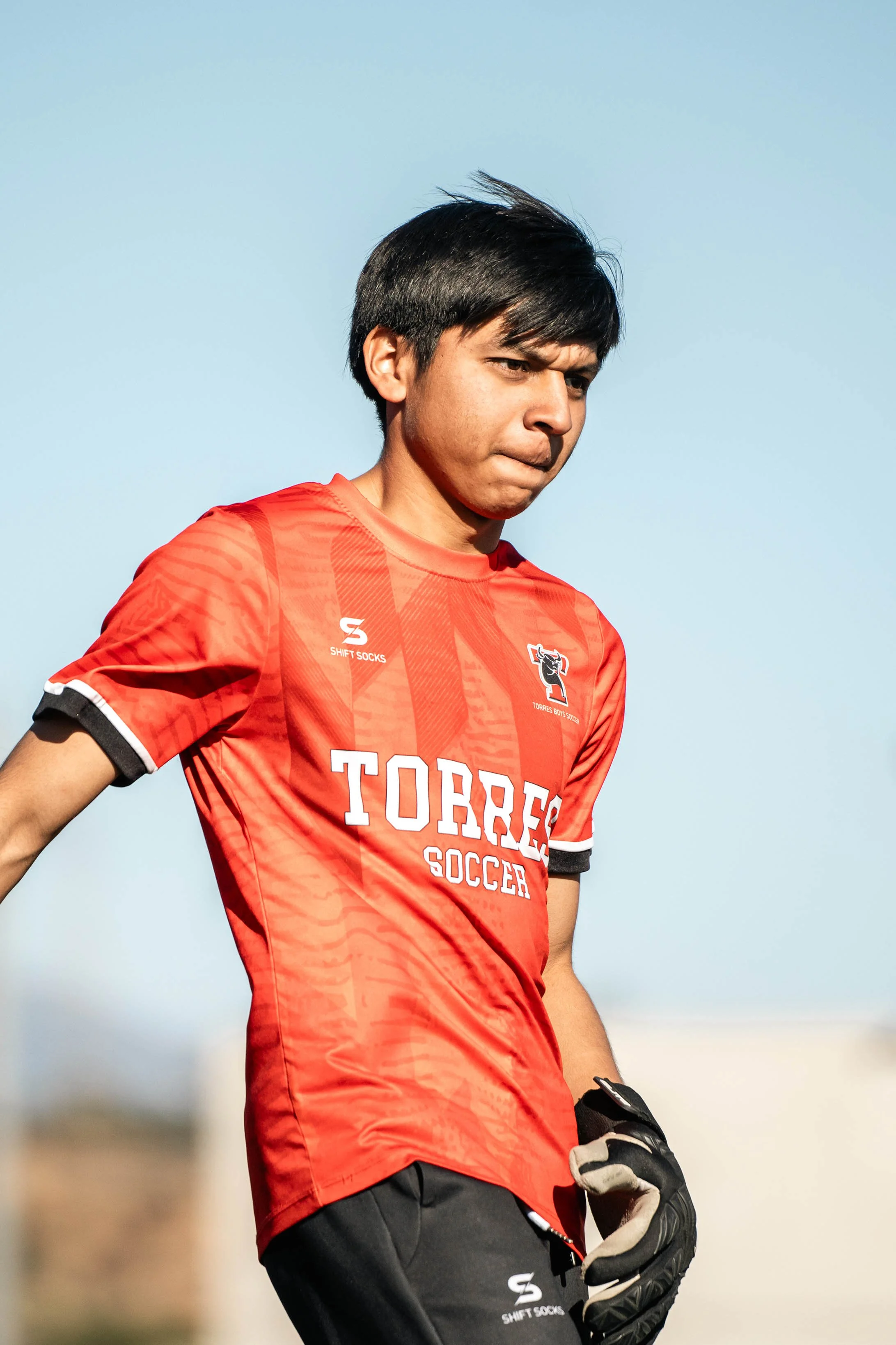 Young soccer player in red jersey with "TORRES SOCCER" text, holding a goalkeeper glove, outdoor setting.