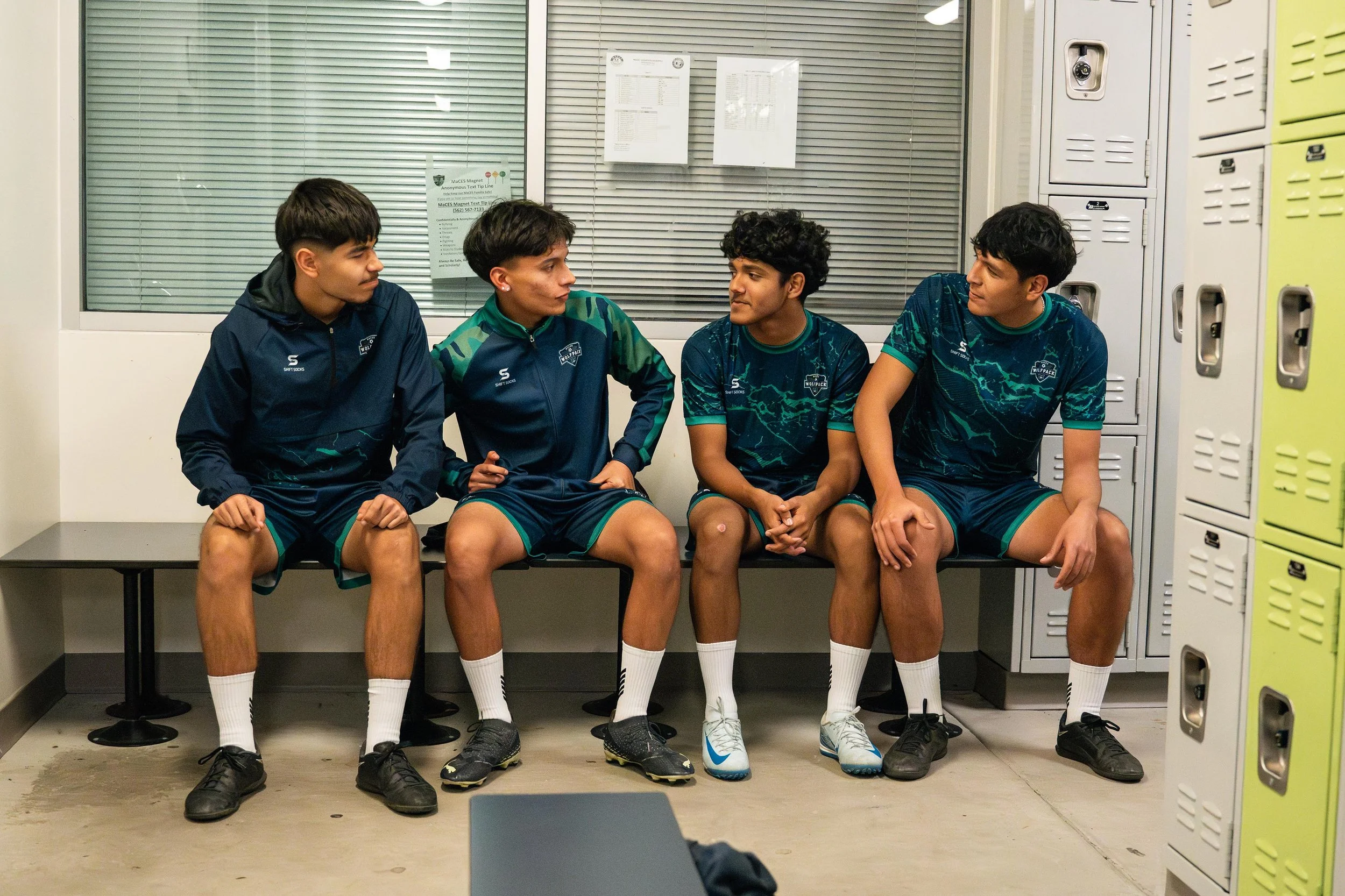 Four young men in sports uniforms sitting in a locker room.