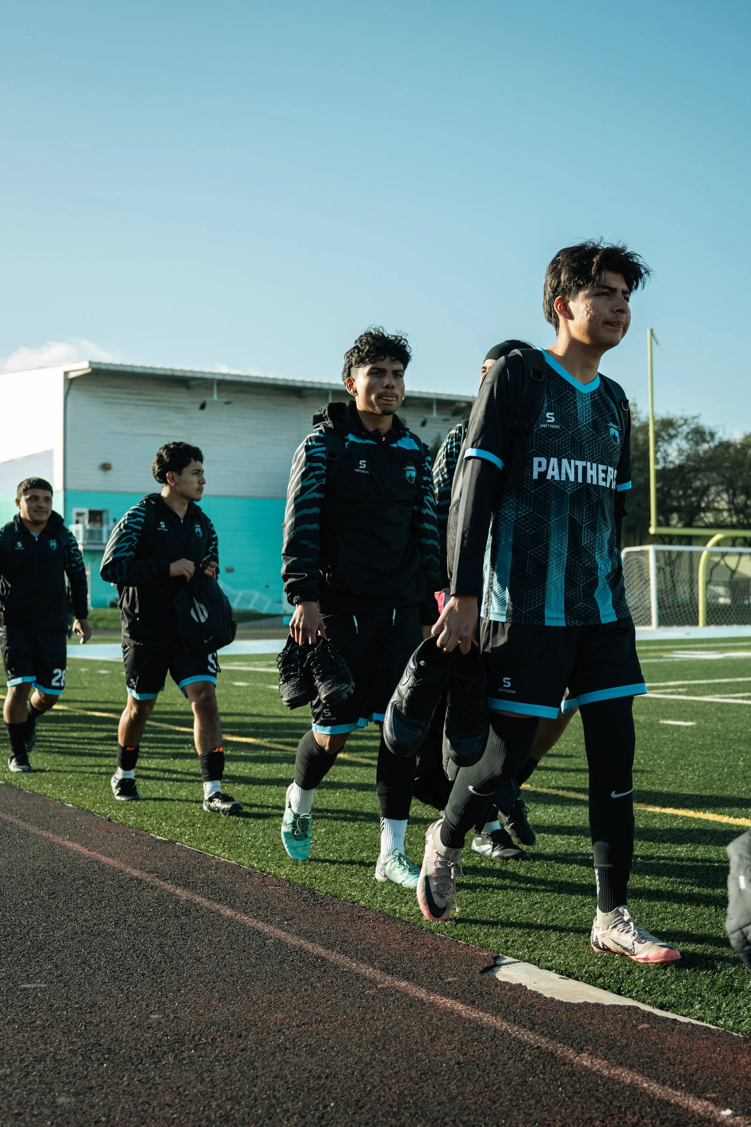 A group of young male athletes walking on a field with their sports gear, wearing black and blue uniforms with 'PANTHER' written on the front, carrying shoes and sports bags.