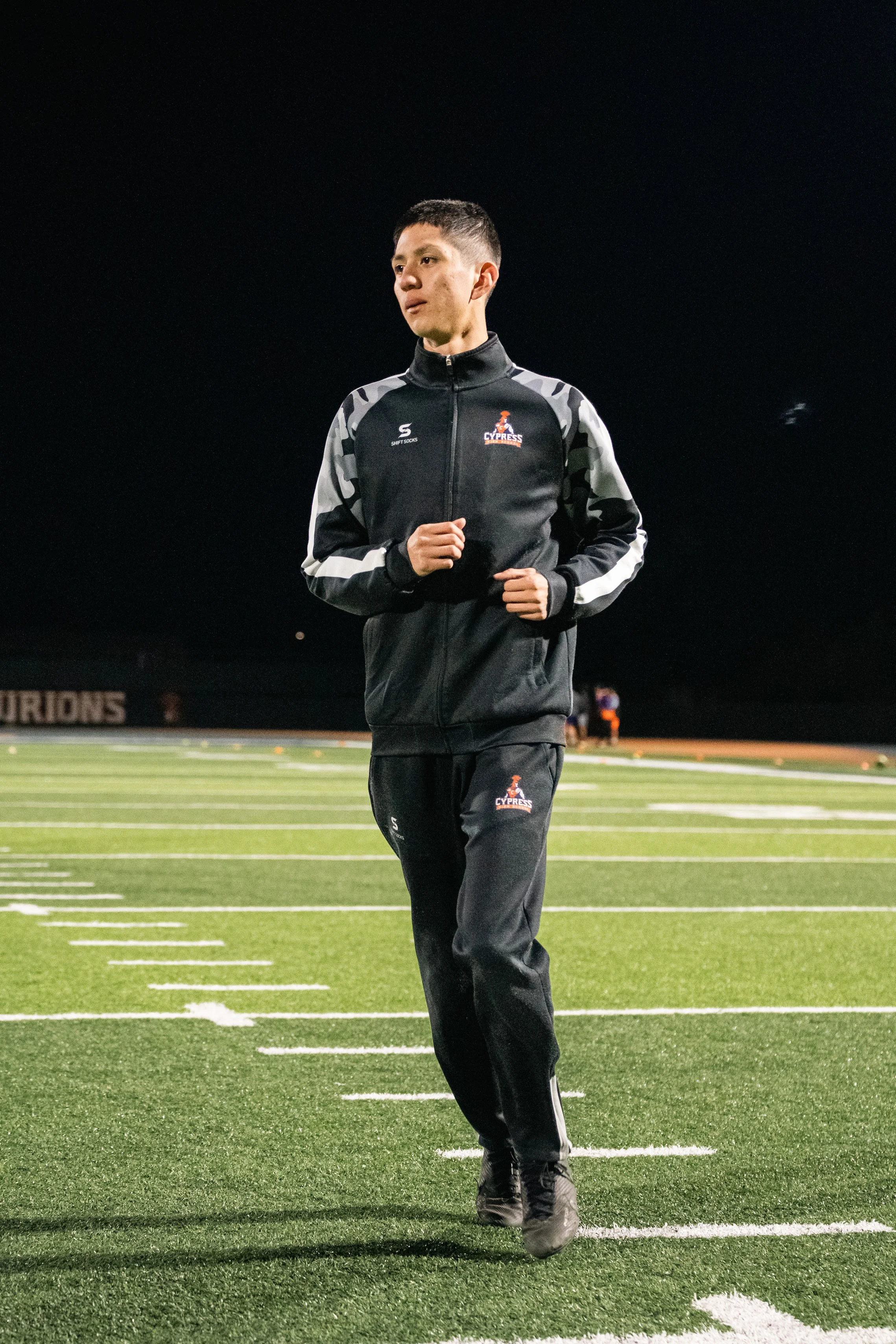 Person jogging on a football field at night, wearing a black sports tracksuit.