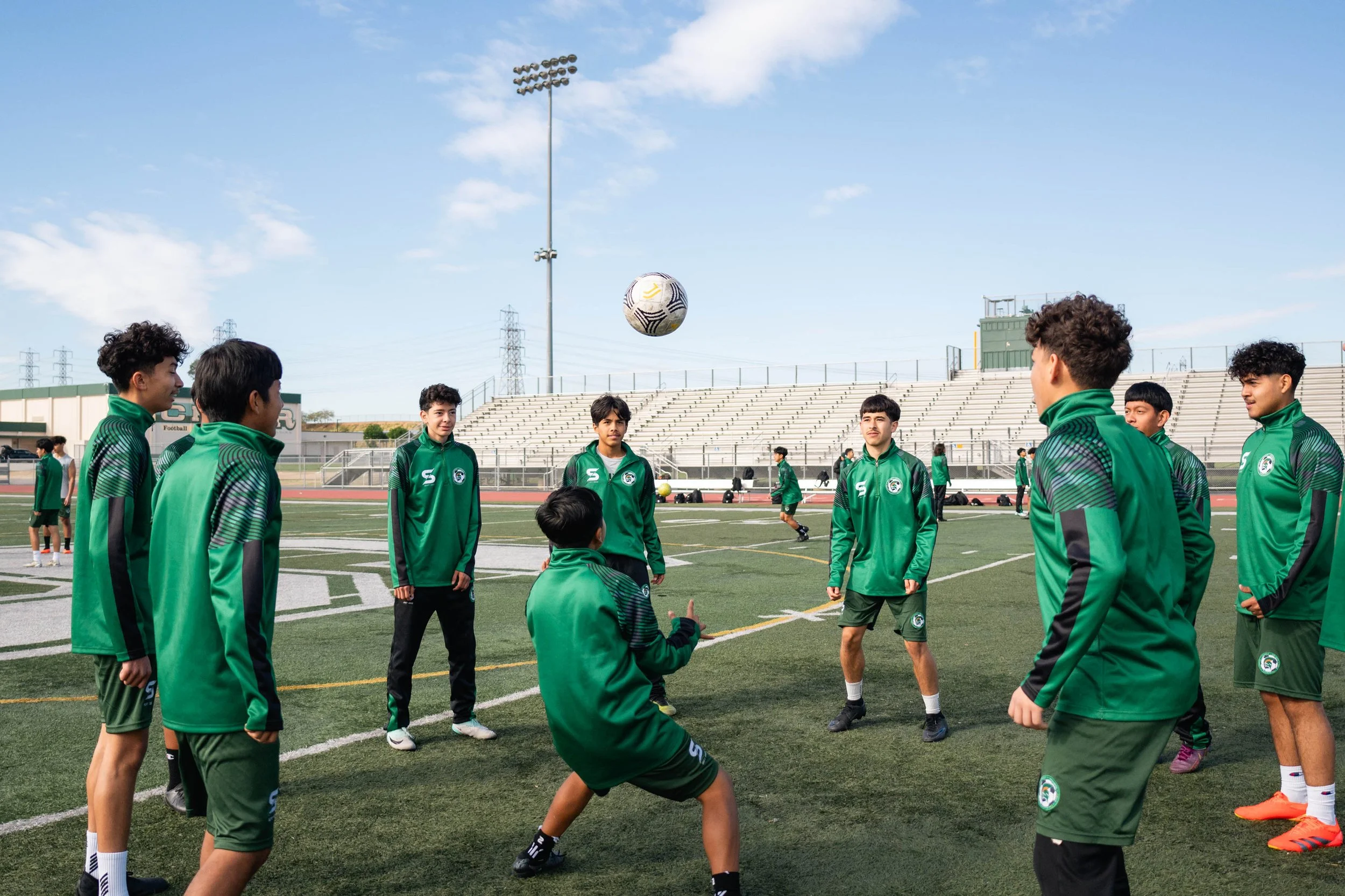 A group of young soccer players in green uniforms practicing on a field, standing in a circle with a soccer ball in the air. The field has a track and empty bleachers in the background, under a clear blue sky.