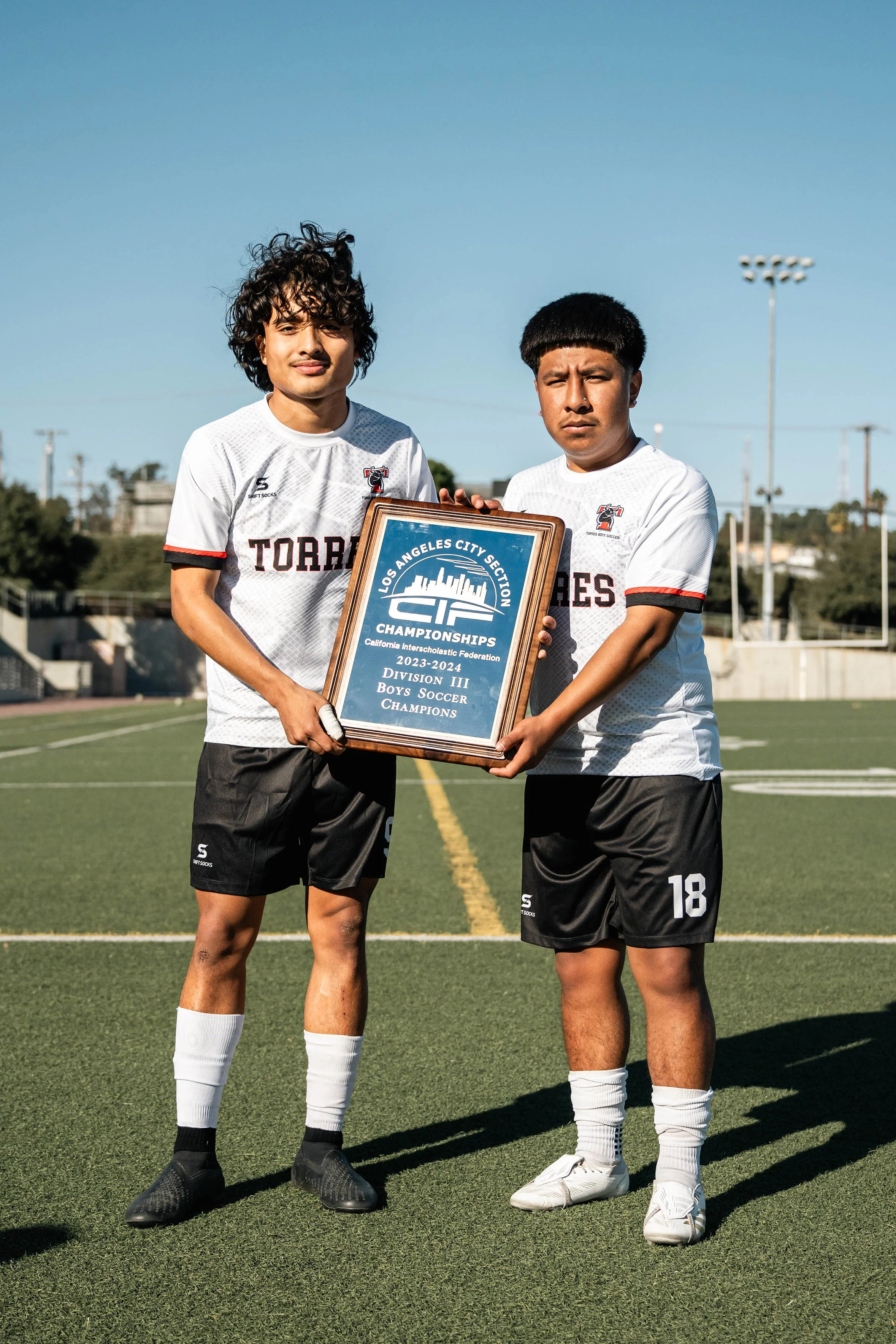 Two soccer players holding a Los Angeles City Section CIF Championship plaque on a field.