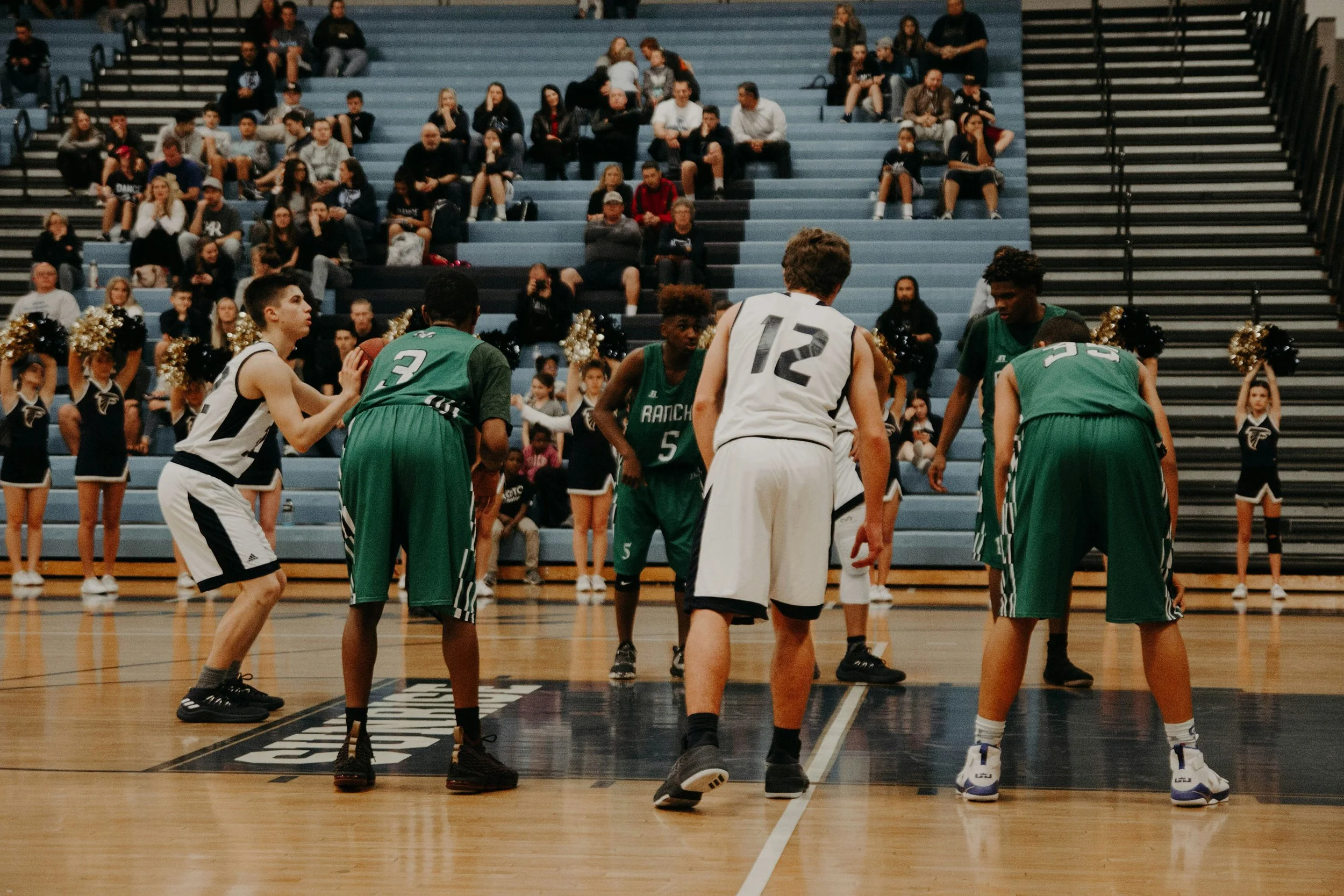 High school basketball game with players in white and green jerseys preparing for a play, surrounded by cheerleaders and spectators in the bleachers.