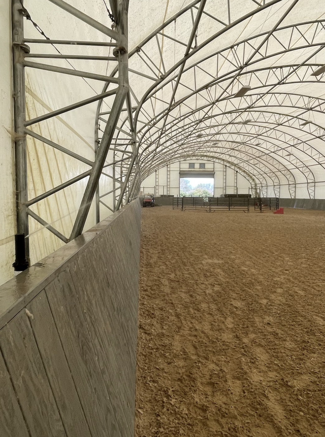Interior of an indoor horse riding arena with metal framework and sand-covered ground.