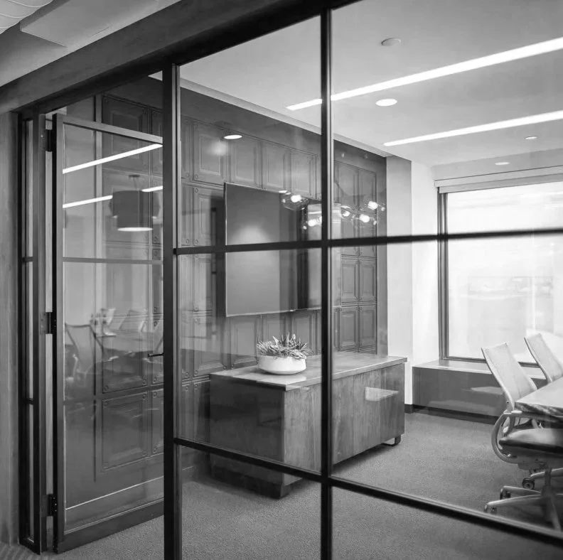 A modern office conference room seen through glass partitions, with a wooden cabinet, potted plant, flat-screen TV, office chairs, and large windows allowing natural light.