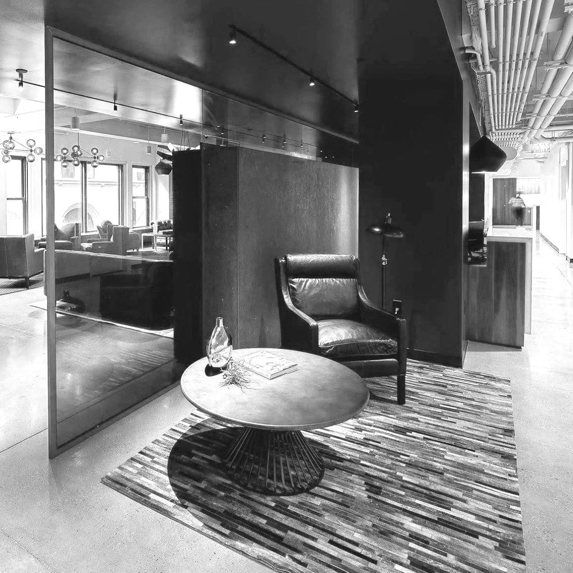 A black and white photo of a cozy corner in a modern lounge or waiting area with a leather armchair, a small round table with a decorative glass vase and a book, a rug, and a lamp, with other seating areas visible through a glass partition.