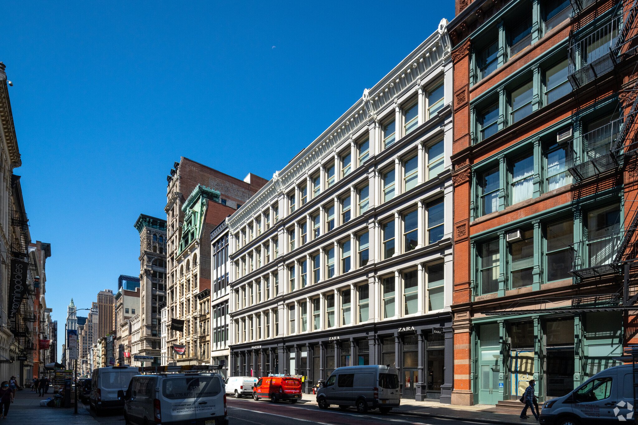 City street with historic buildings, storefronts including Zara, cars parked along the curb, pedestrians walking, under a clear blue sky.