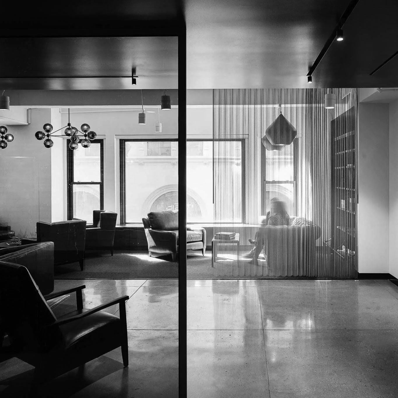 A black and white photo of a modern interior space separated by a glass wall. On one side, there are chairs and a lamp. On the other side, a woman sitting on a sofa near large windows, with curtains, and a city view outside.