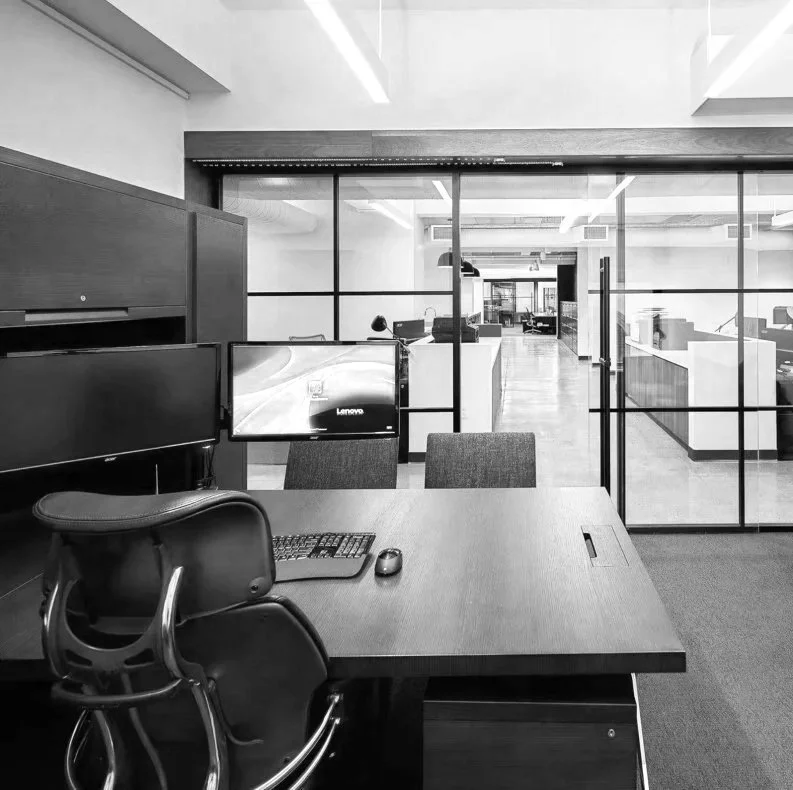 Modern office workspace with a desk, ergonomic chair, two computer monitors, and a glass partition separating it from additional workstations in the background.