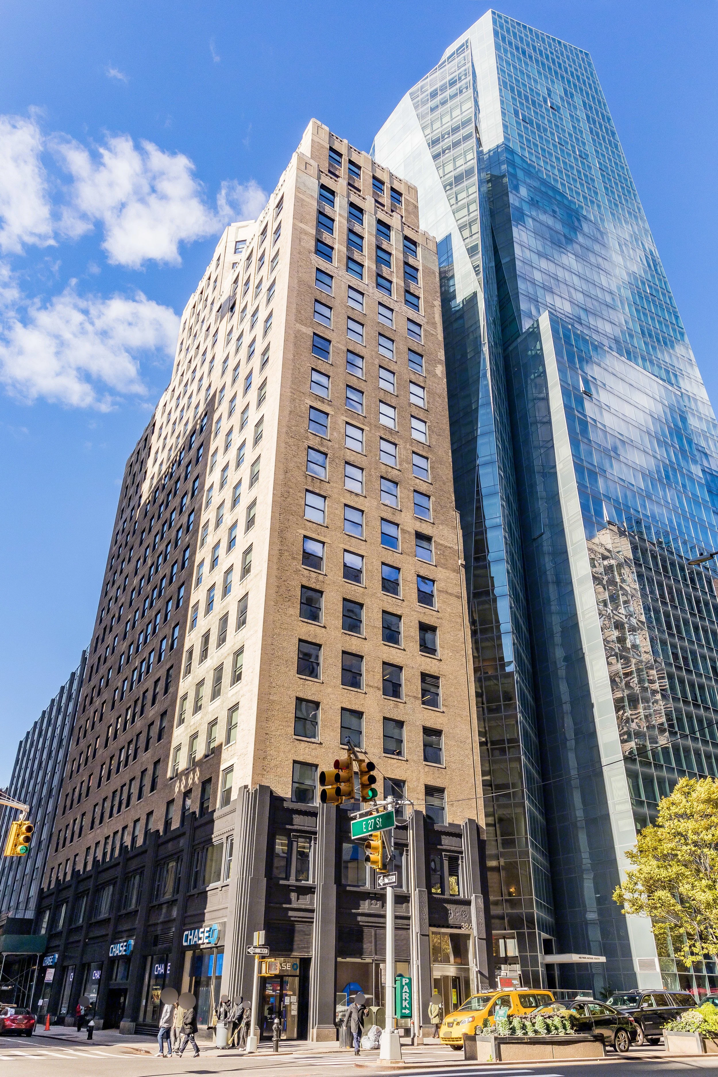 386 Park Avenue South Facade - View of tall city buildings, including a historic stone building and a modern glass skyscraper, with people crossing the street and cars parked at an intersection.