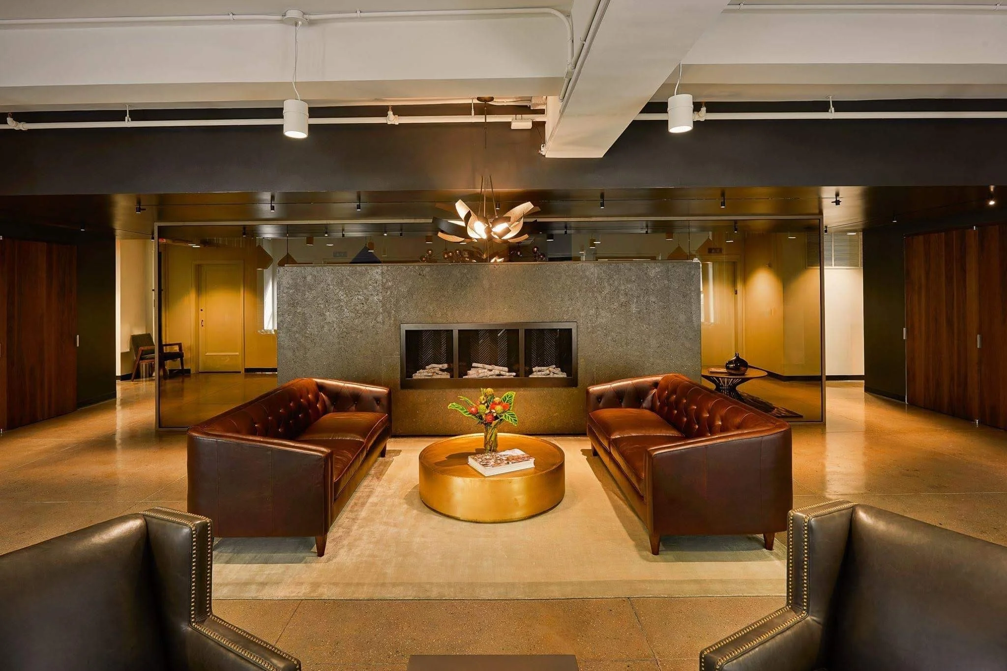 A modern lobby with two brown leather sofas facing each other, a round gold coffee table with a flower arrangement, and a fireplace with stacked logs, set against a dark textured wall. There are wall-mounted lights and a contemporary chandelier hanging from the ceiling.
