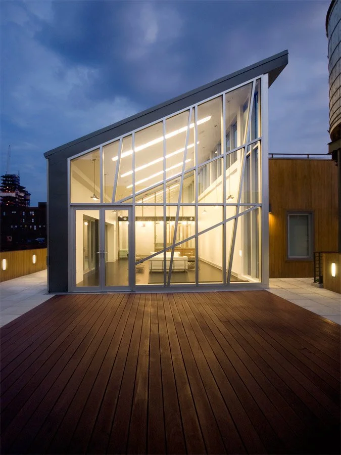 503-511 Broadway Penthouse - Modern house with large glass windows and an angular roof, illuminated from inside, on a rooftop patio with wooden flooring at dusk.