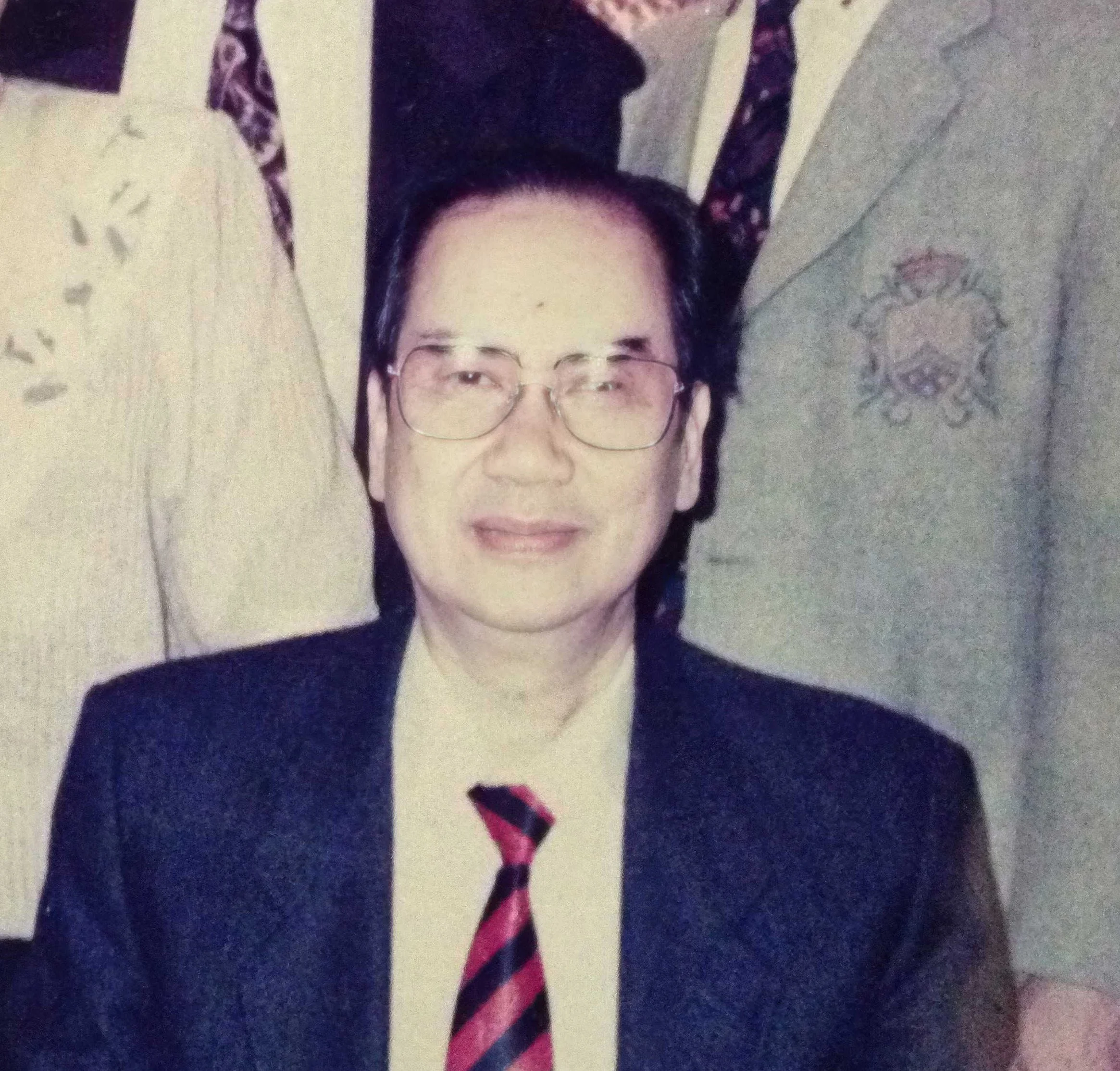 Founder - A middle-aged man with glasses in a black suit, white shirt, and red striped tie, standing in front of patterned and solid-colored fabrics.