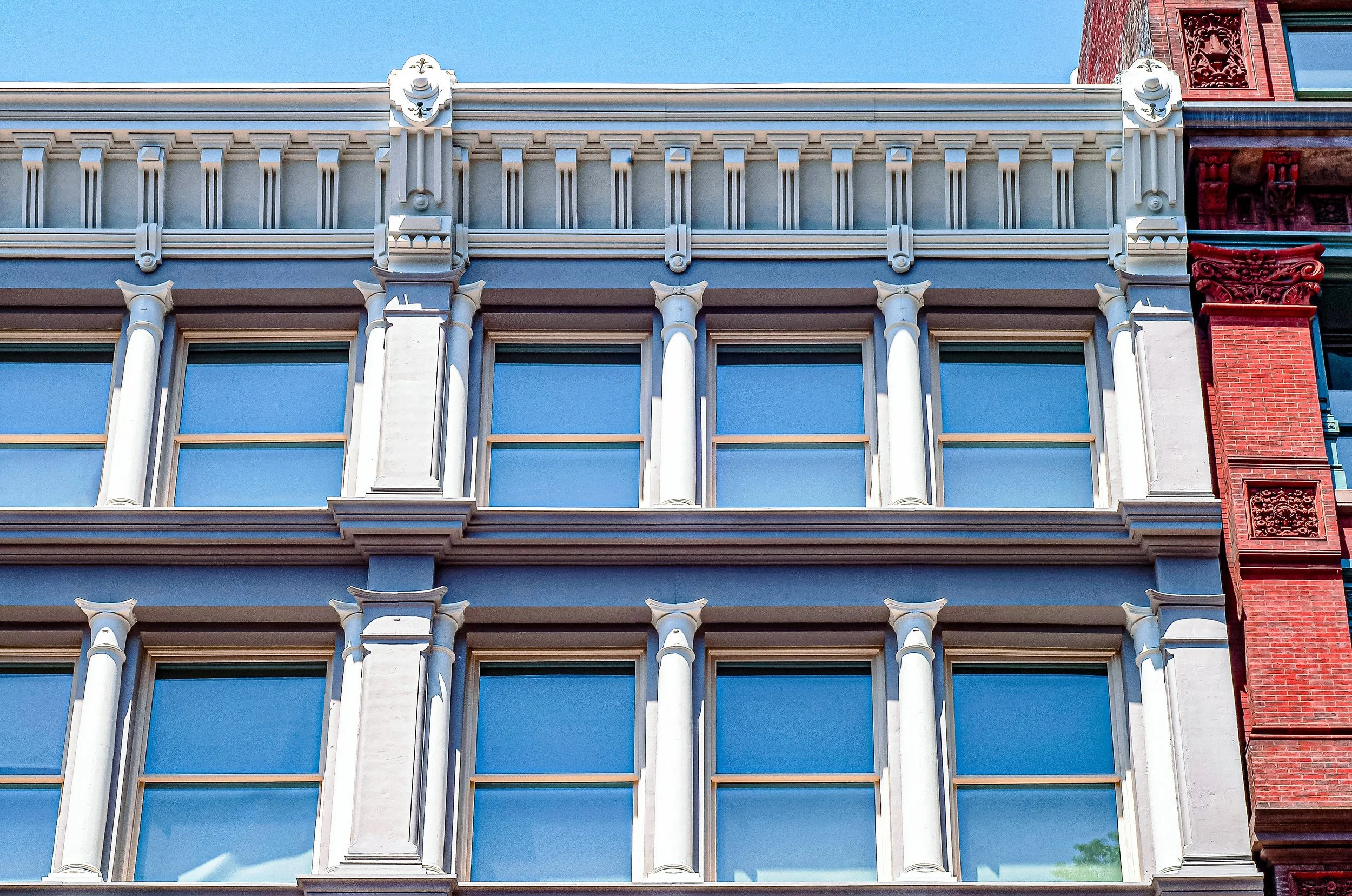 Close-up of an ornate building facade with three floors, displaying decorative architectural details like columns, corbels, and window moldings. The building has white elements and large blue-tinted windows under a clear blue sky. SoHo cast-iron.