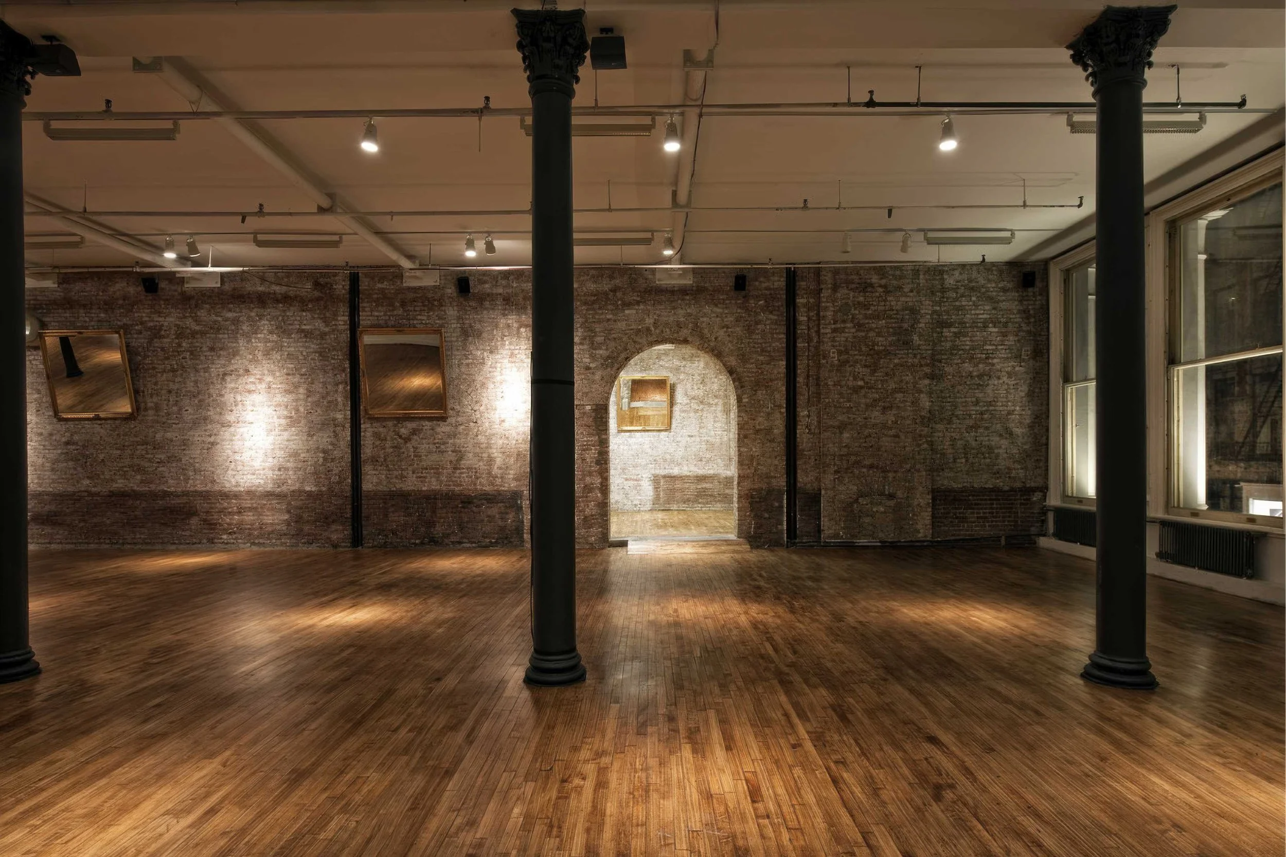 503-511 Broadway Interior - An empty art gallery with exposed brick walls, polished wooden floors, large windows on the right, and black columns. Two framed mirrors hang on the back wall, and a brick archway leads to another room.