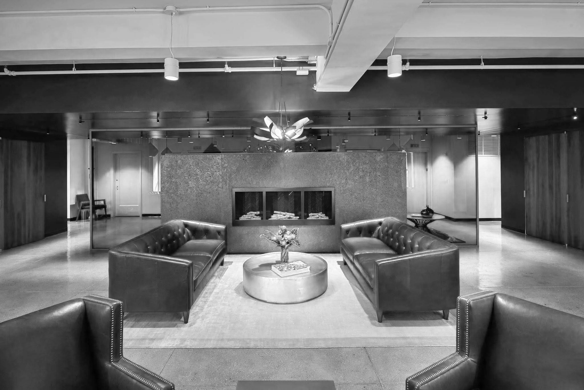 Black and white photo of a modern lobby with two black leather sofas facing each other, a circular coffee table with a decorative flower arrangement in the center, a textured wall behind the sofas, and a fireplace feature. There are chairs and a door in the background, and ceiling lights are visible.