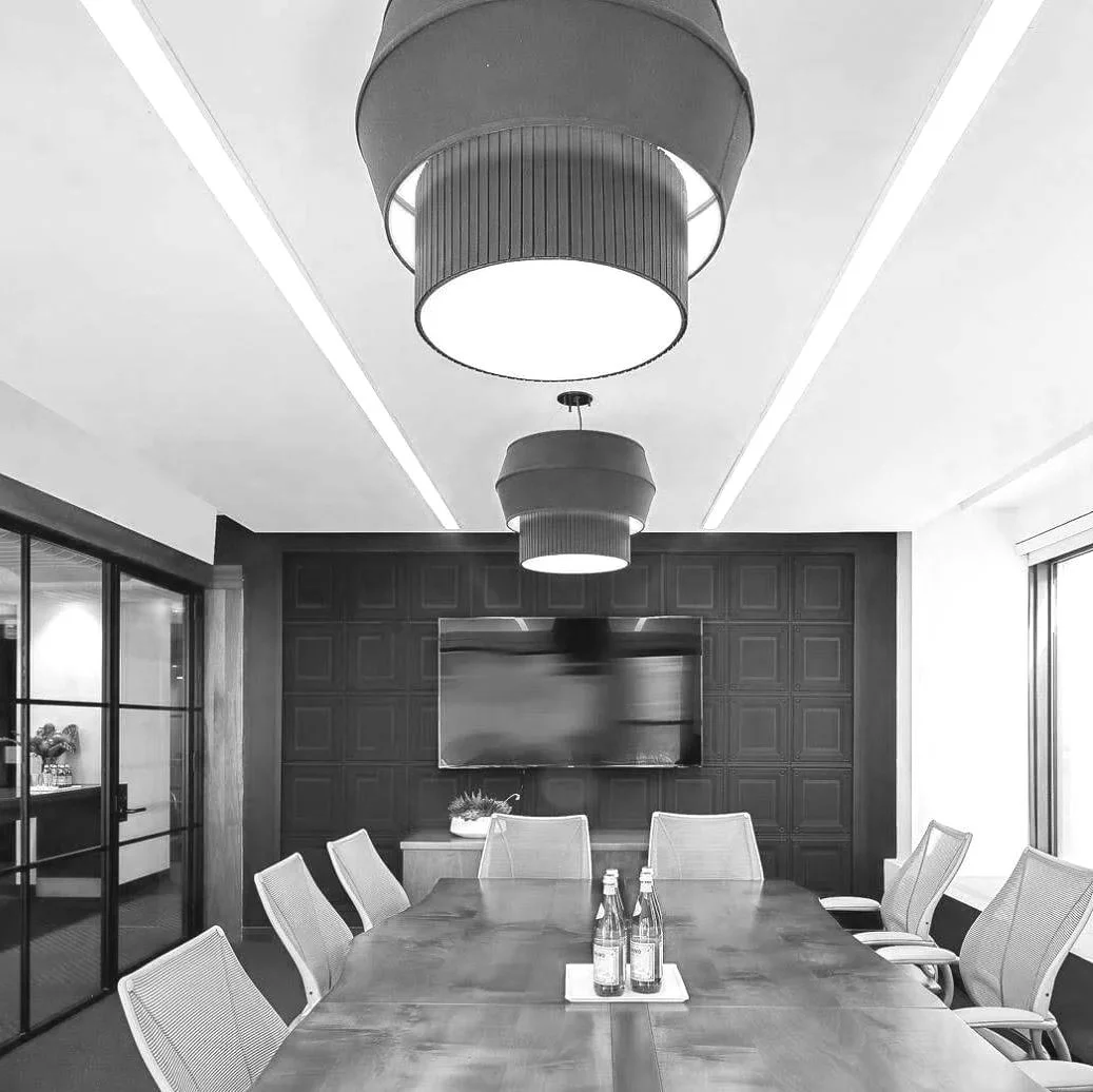 A modern conference room with a large wooden table, eight mesh chairs, a wall-mounted TV, dark wood paneling, and two large pendant lights hanging from the ceiling.