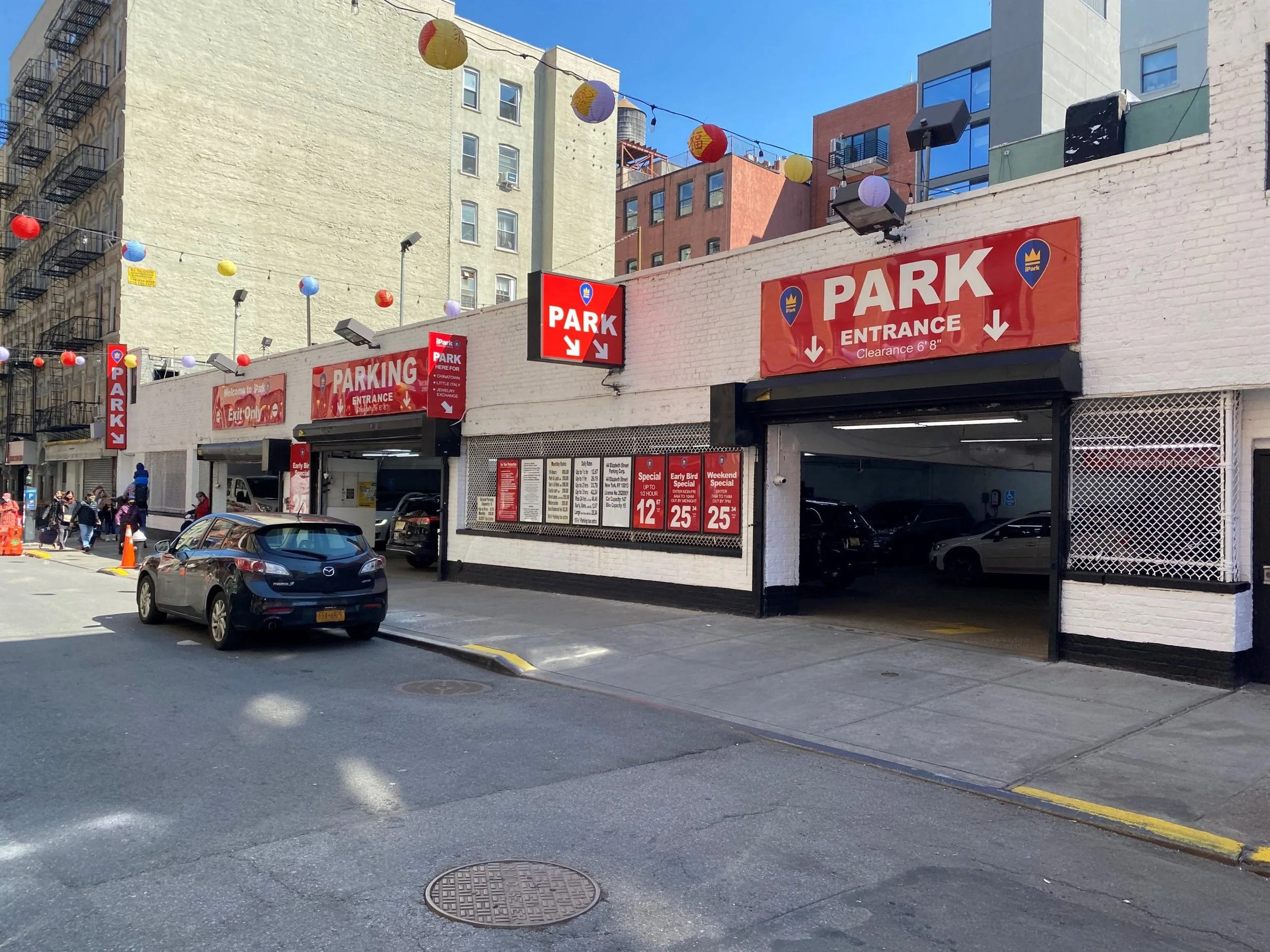 52 Elizabeth St. Facade - Entrance to a parking garage with red signs labeled 'PARK' and 'ENTRANCE'. Several cars parked inside the garage, with a few cars and pedestrians outside on the street. Colorful lanterns hanging overhead.