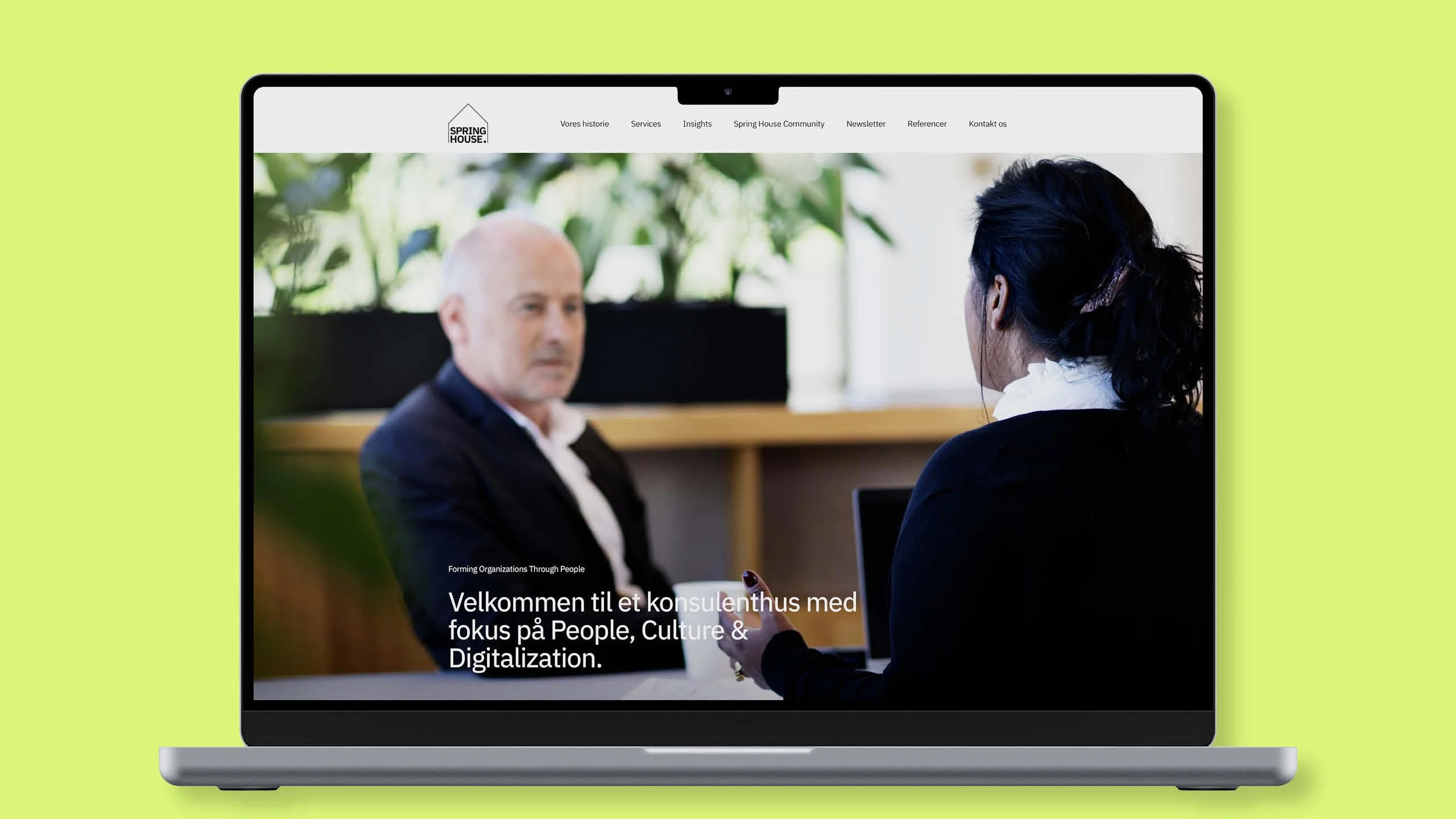 Laptop displaying a website with a photo of a man and a woman in a professional setting. The website header includes menu options and a logo that reads "Spring House." The text on the website is in Danish and mentions welcoming to a consultancy focused on People, Culture, and Digitalization.