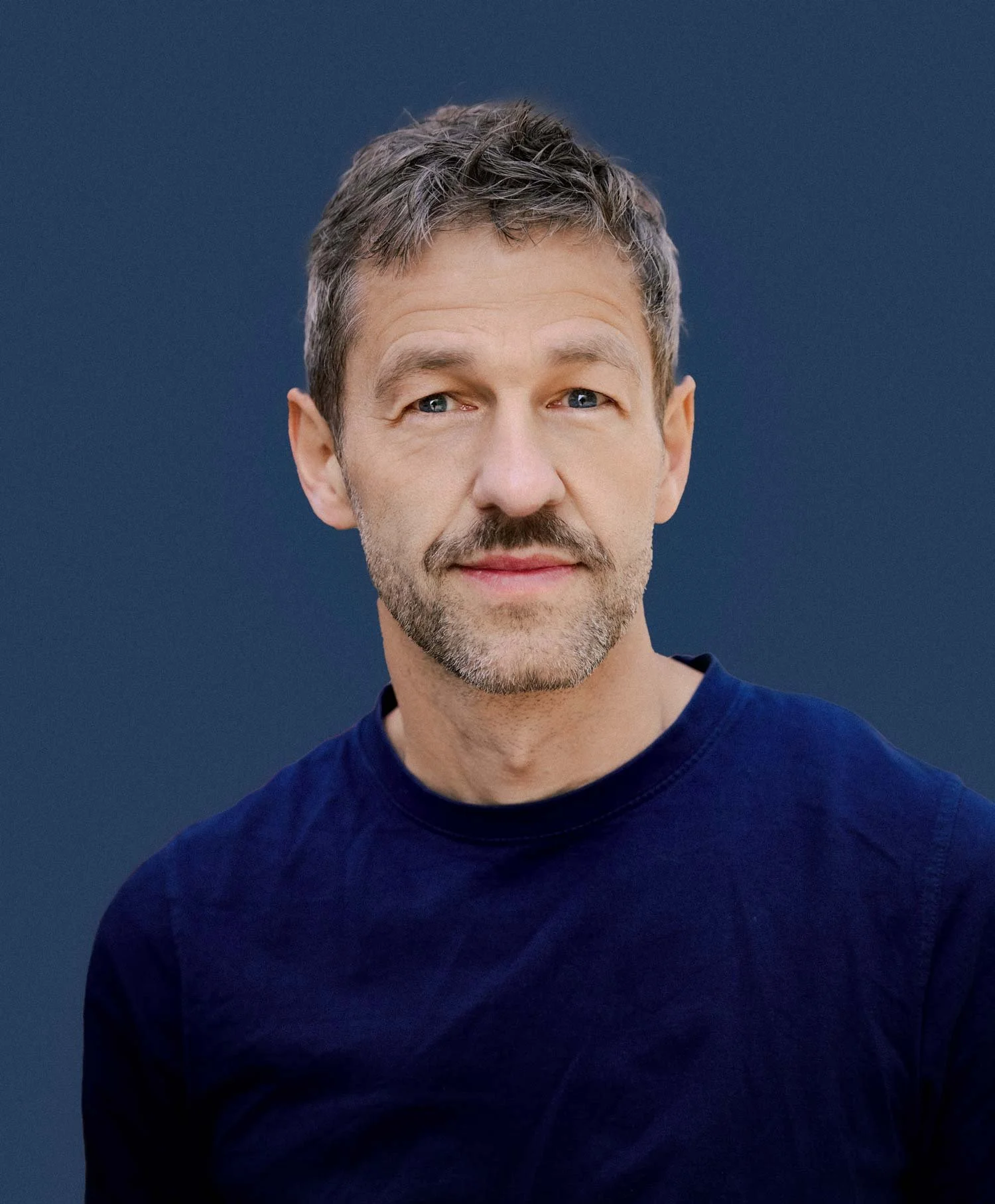 Close-up portrait of middle-aged man with gray hair and beard, wearing a navy blue shirt, against a dark blue background.
